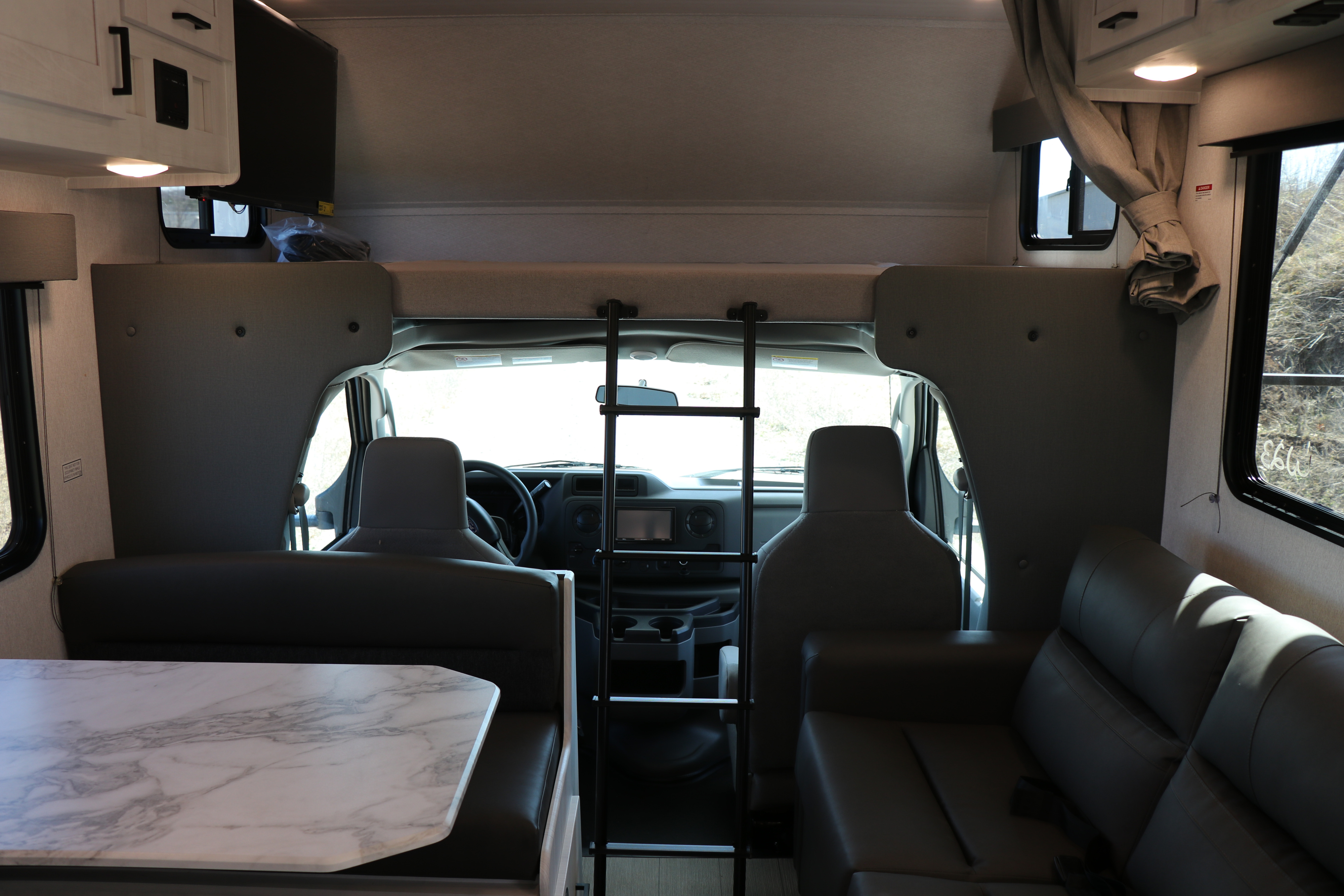 RV rental interior view: dinette with table, front cab area with steering wheel, overhead bunk bed with ladder, and sofa.. East to West entrada 2025