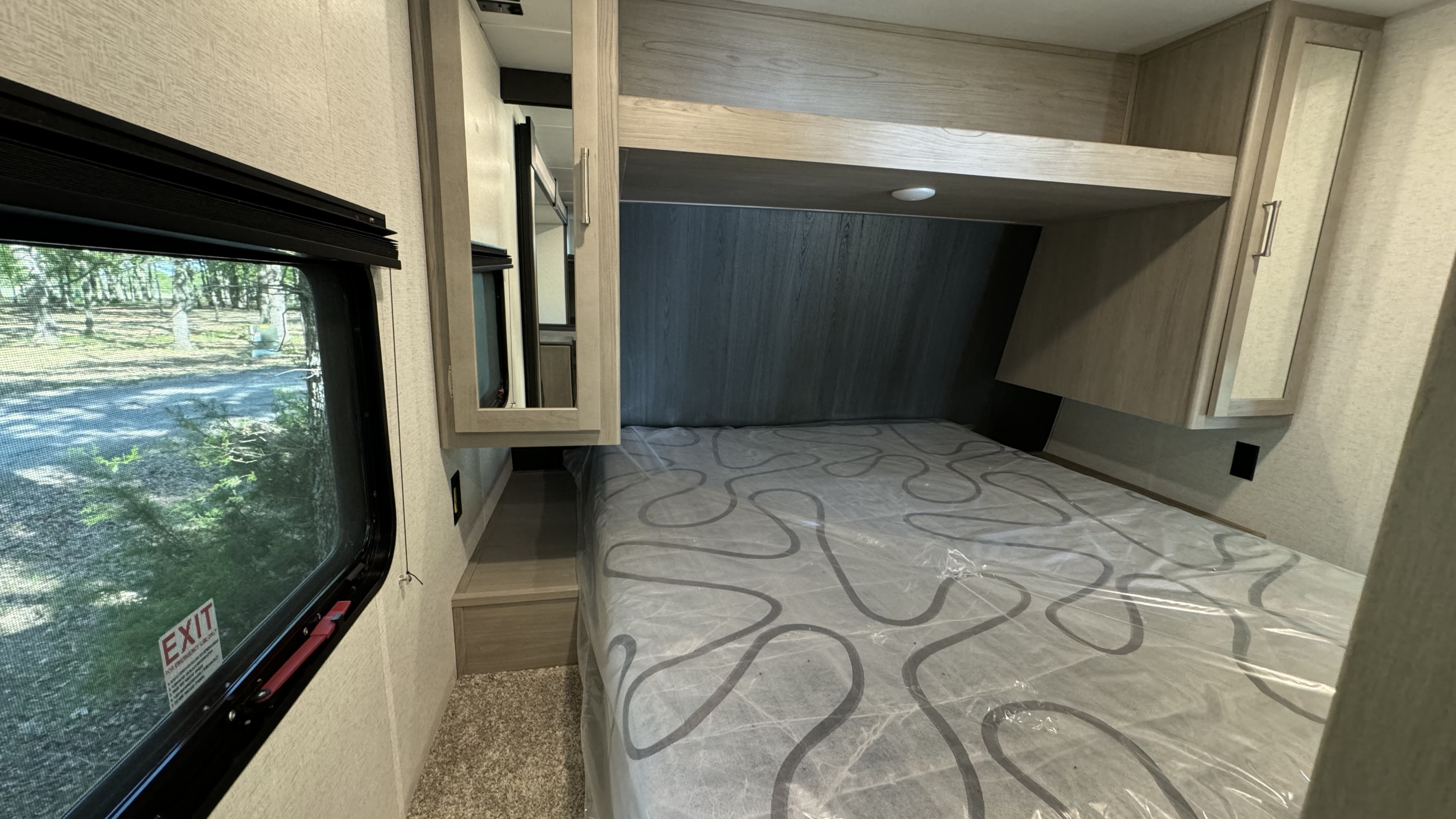 Cozy rv rental bedroom showing a bed with plastic-wrapped mattress, light wood overhead cabinets, and an emergency exit window.. Springdale 261BHC 2024