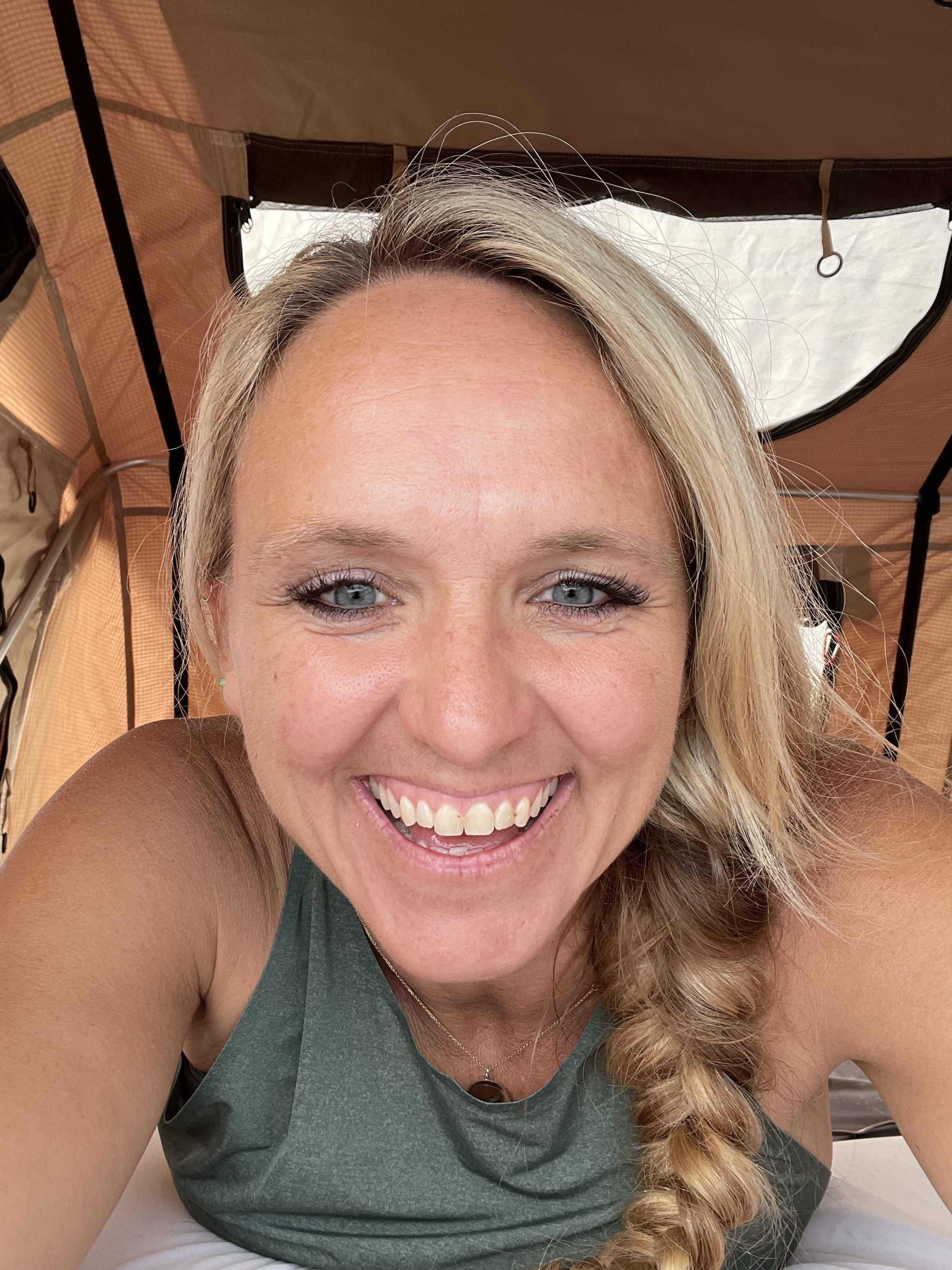 A happy blonde woman with blue eyes smiles broadly, cozy inside an RV rental with fabric walls. Ready for camping adventures!. SPCNS Other 2020