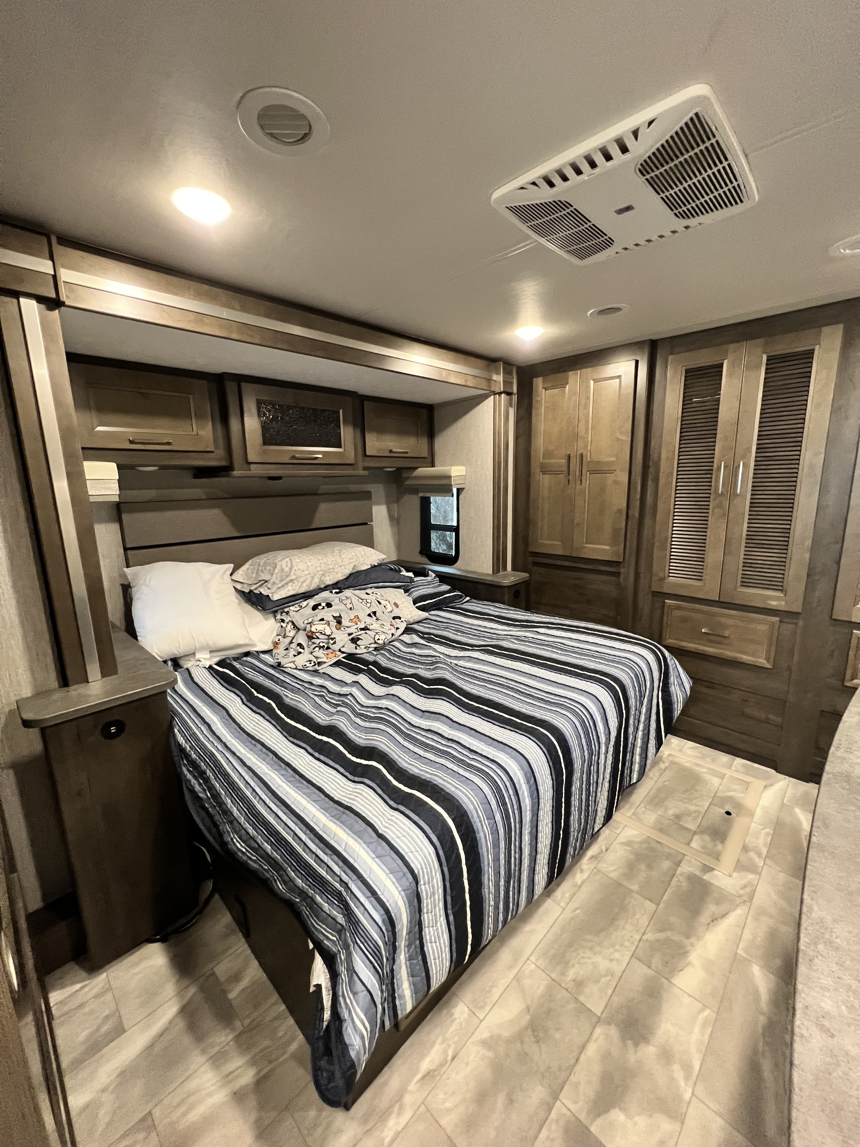 A cozy RV rental bedroom, featuring a large bed with a blue and white striped comforter, wooden cabinets, and a ceiling AC unit.. Forest River Coachmen Sportscoach SRS 2021