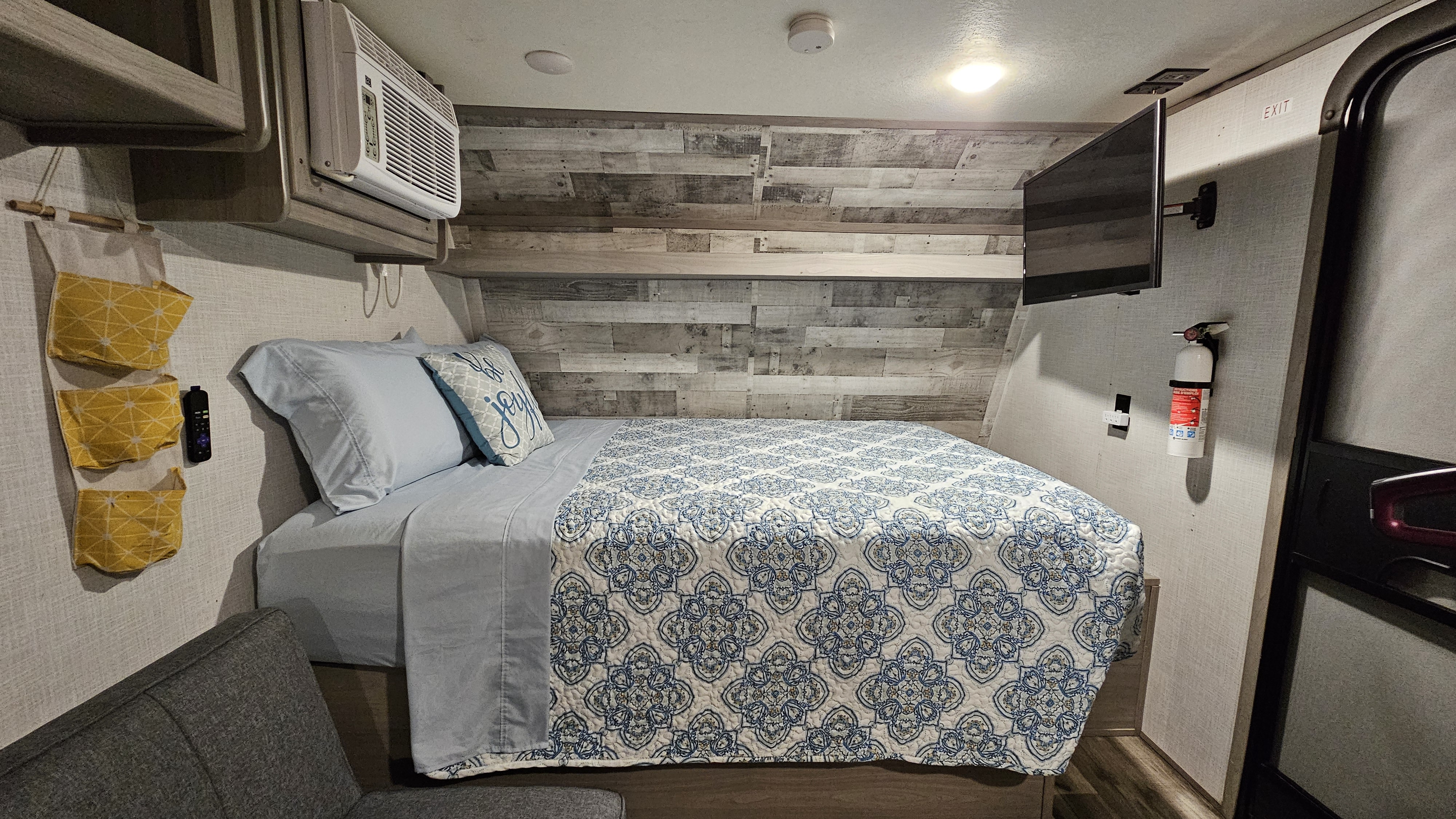 Cozy RV rental bedroom featuring a patterned bed, wall-mounted TV, AC, and distressed wood accent wall.. Coleman Lantern 17B 2023