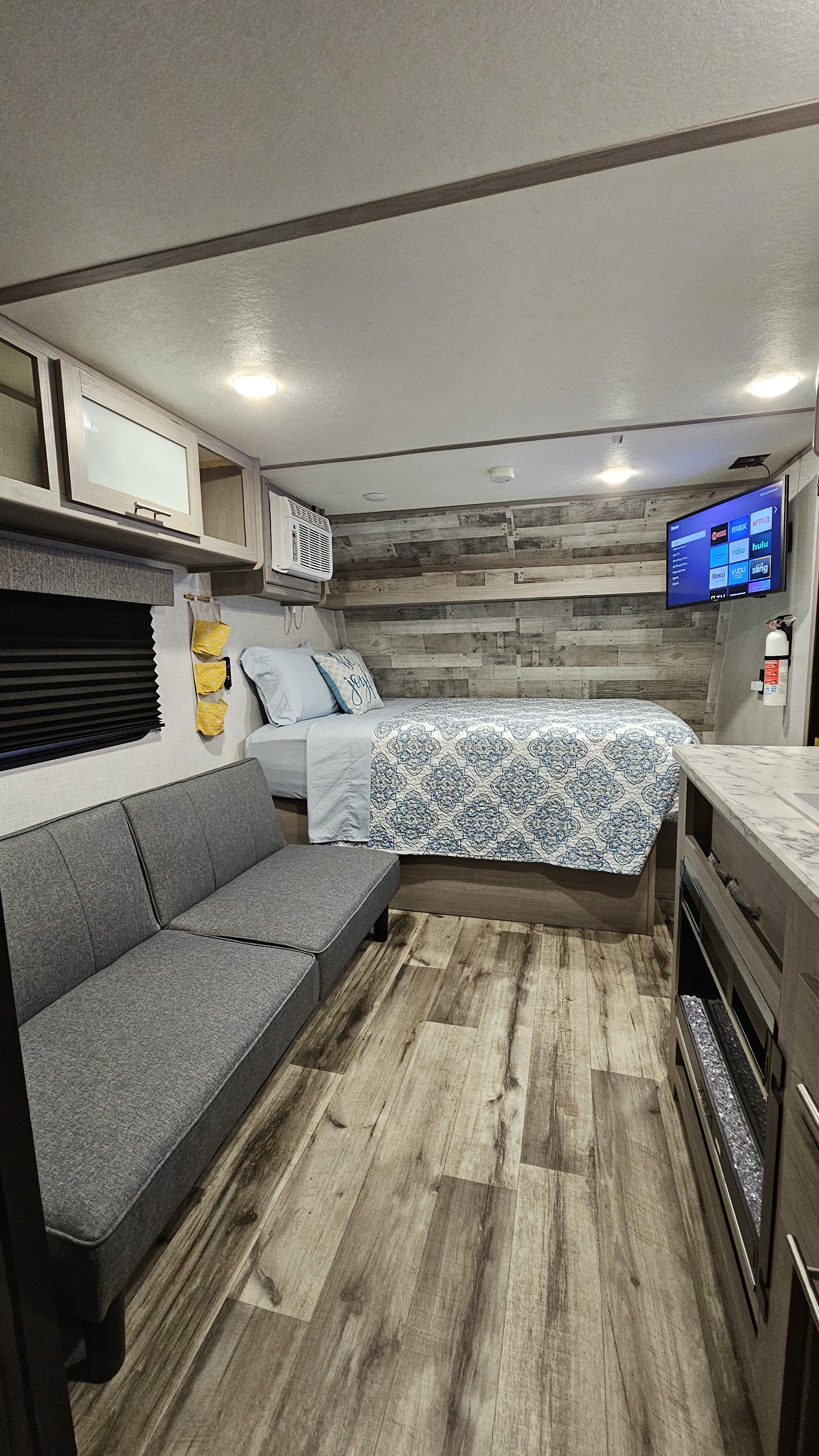 Cozy RV rental interior with a bed, gray futon, wood-look floor, plank accent wall, and entertainment TV.. Coleman Lantern 17B 2023