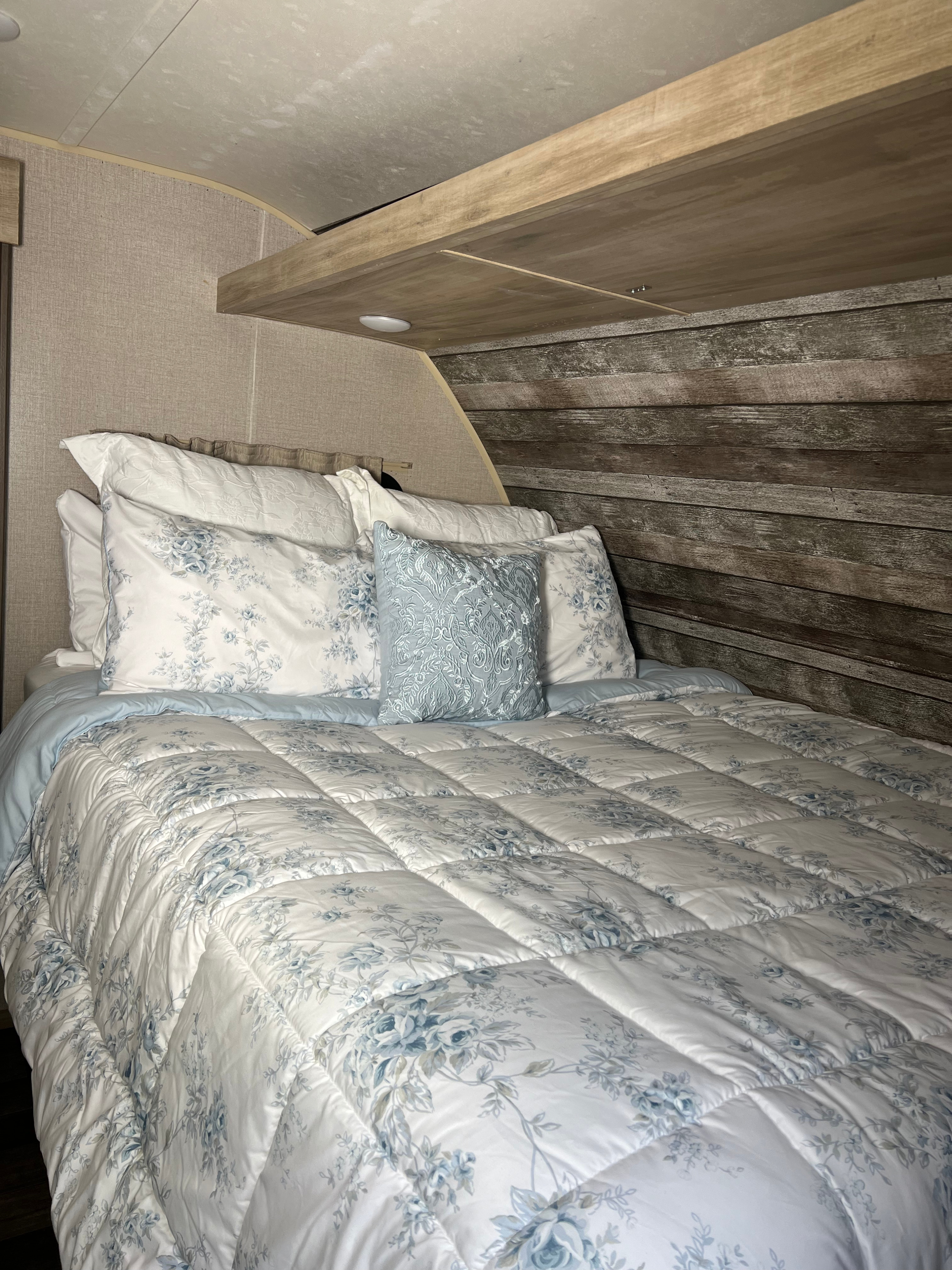 Cozy RV rental bedroom featuring blue floral bedding, a rustic wood headboard, wood shelving, and built-in lighting.. Coachmen Catalina Summit 2021