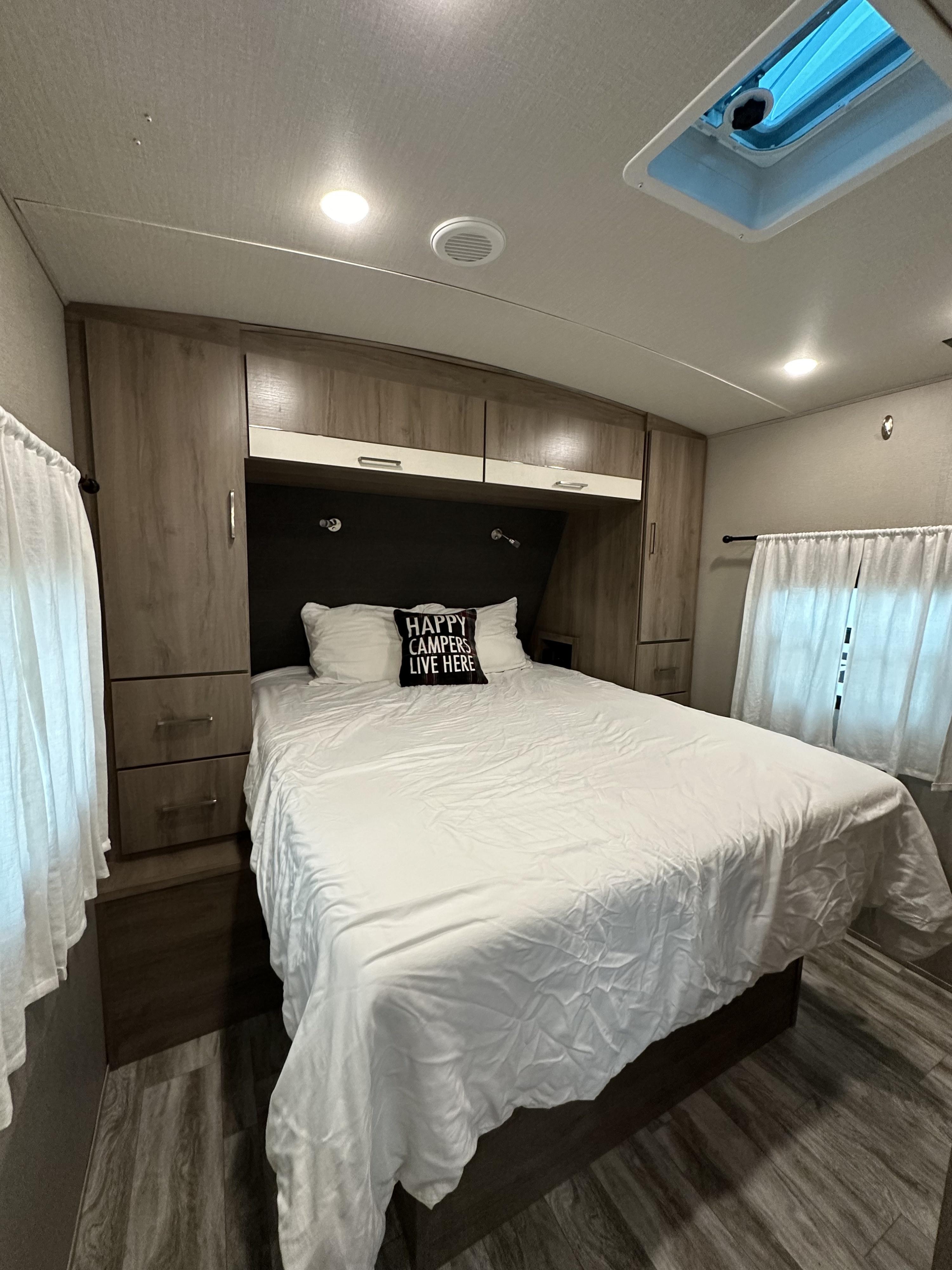 Cozy RV rental bedroom featuring white bedding, "Happy Campers" pillow, built-in wooden cabinetry, and a skylight for natural light.. Grand Design Imagine 2022