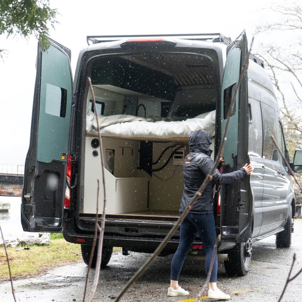 Person by an open dark camper van in light snow, revealing the interior bed. Explore van life with this RV rental option.. Ford Transit 2022