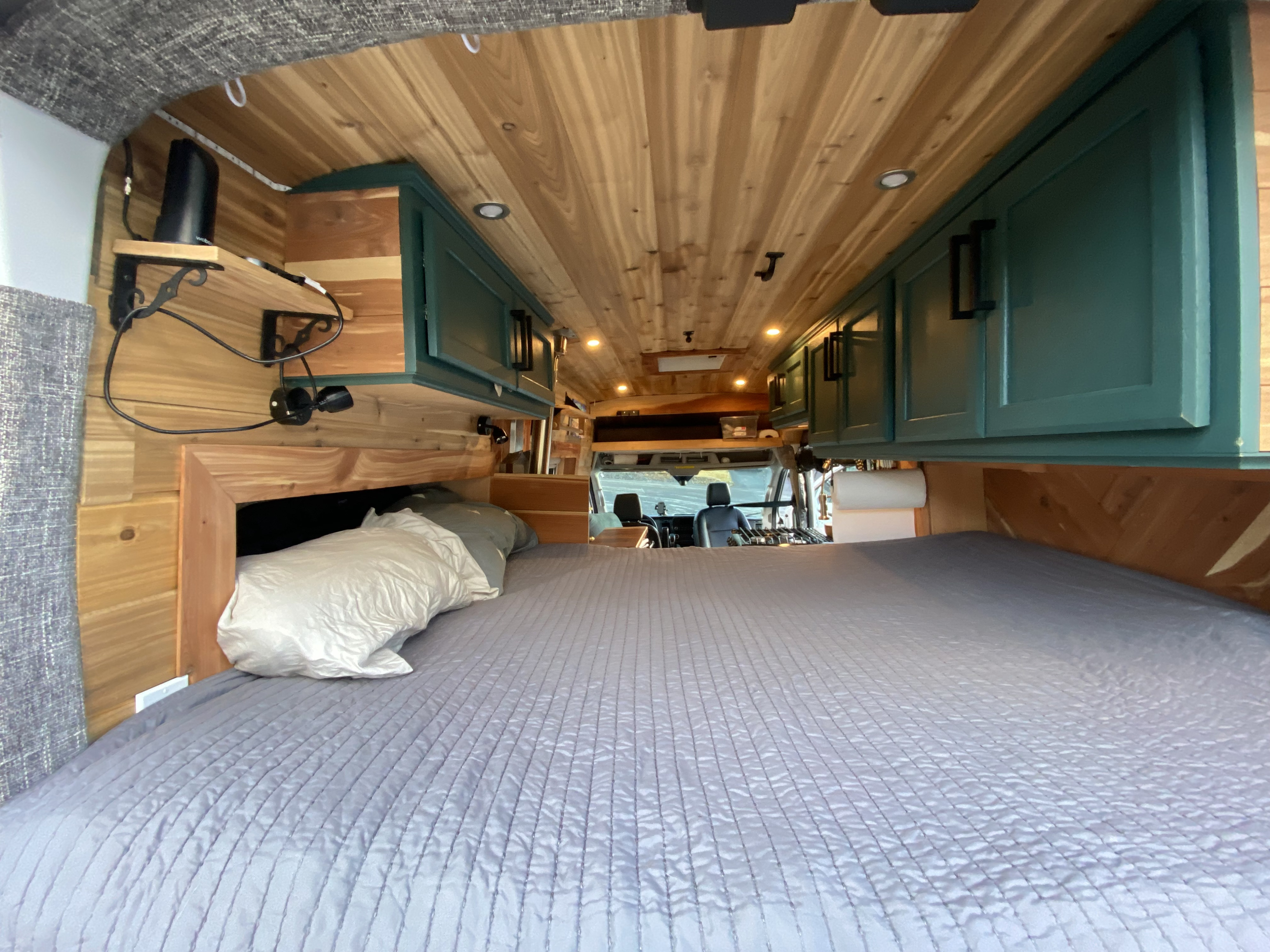 An RV rental's inviting sleeping area featuring a quilted bed, warm wood paneling, and teal overhead storage cabinets.. Ford Transit 2020