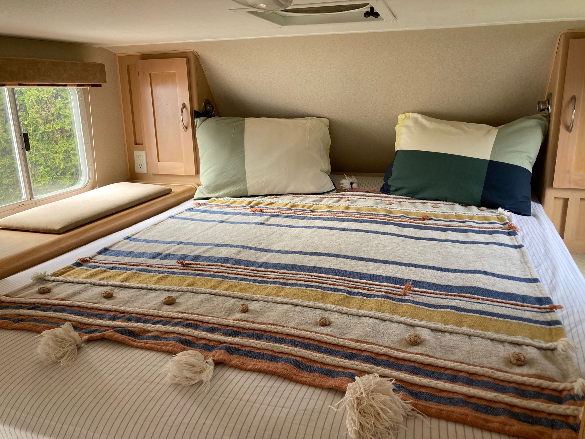 An RV rental bedroom featuring a striped bedspread in blue, yellow, and orange, colorful pillows, and a window view.. Chevrolet Other 2010
