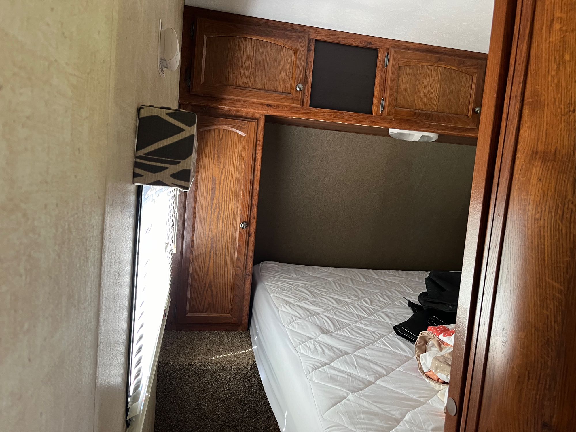 An RV rental bedroom with a white bed, wood overhead cabinets, and a window with blinds.. Keystone RV Hideout 2015