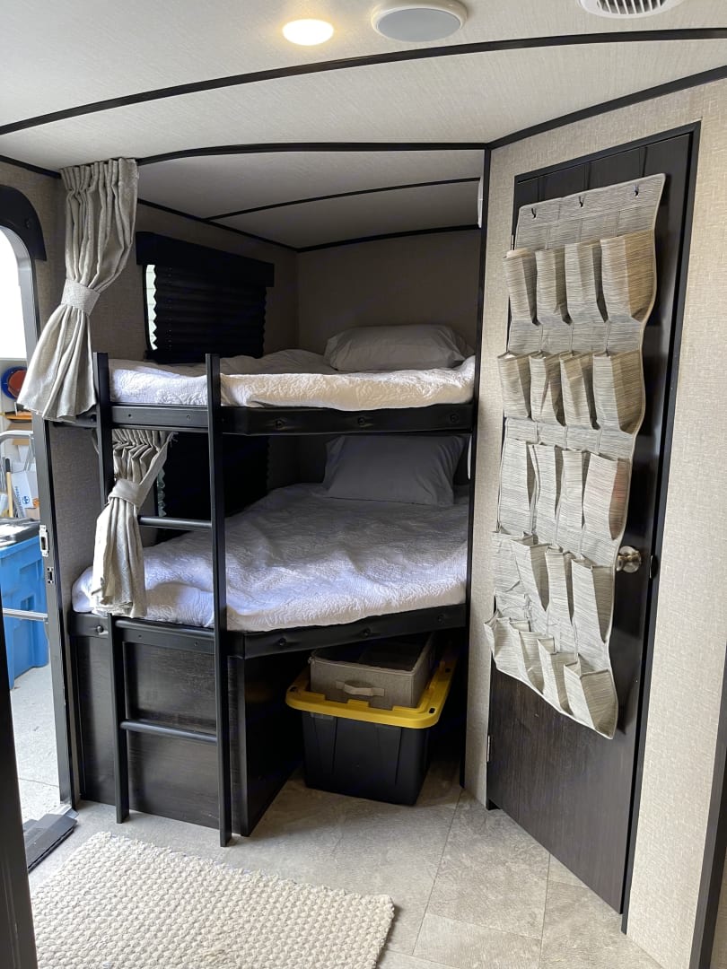 An RV rental interior featuring stacked bunk beds with white bedding, storage bins, and a handy door-mounted shoe organizer.. Grand Design Imagine 2019