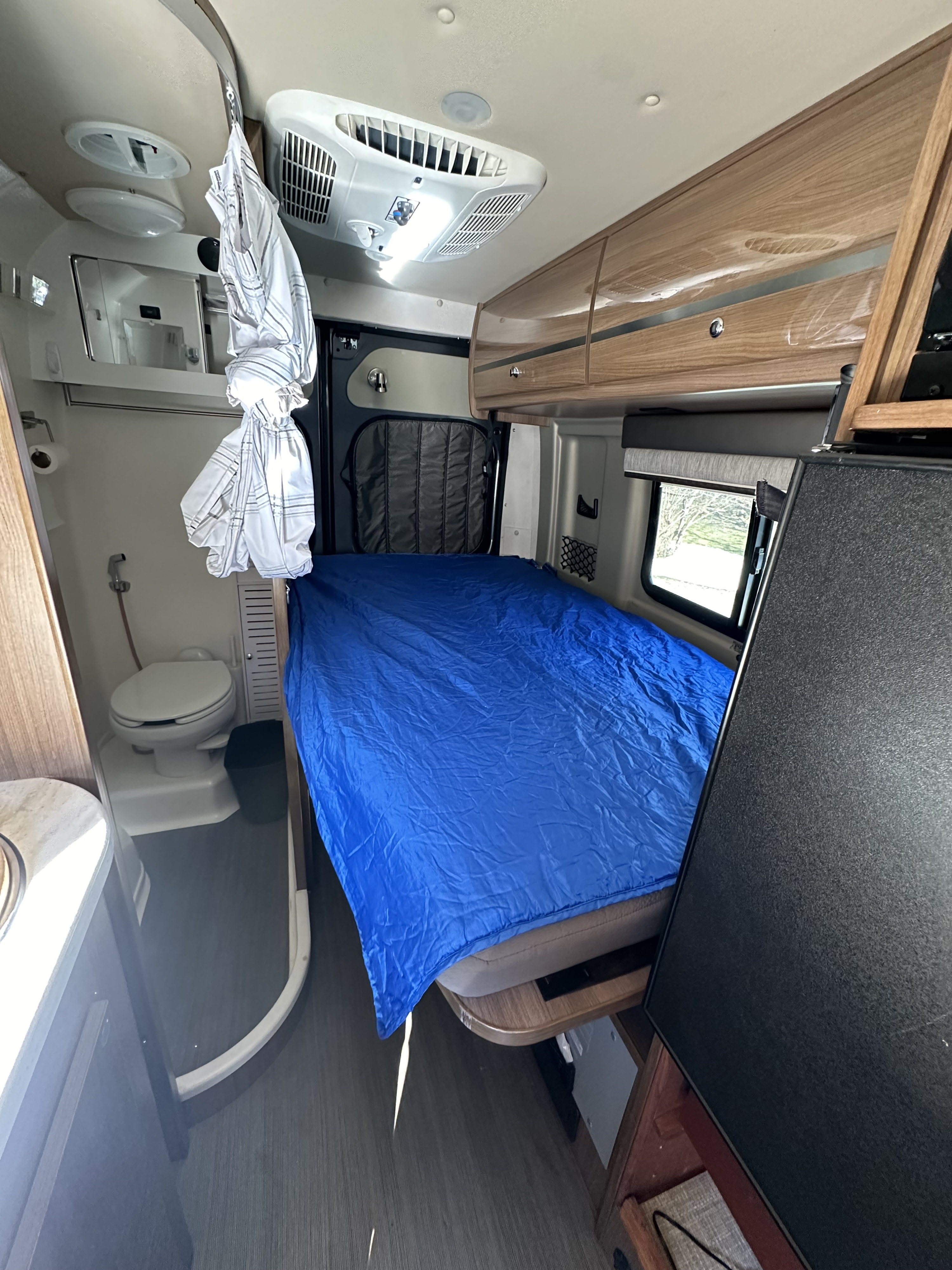 Compact rv rental interior with a blue bed, small bathroom, and overhead storage, ideal for mobile living.. Winnebago Travato 2016
