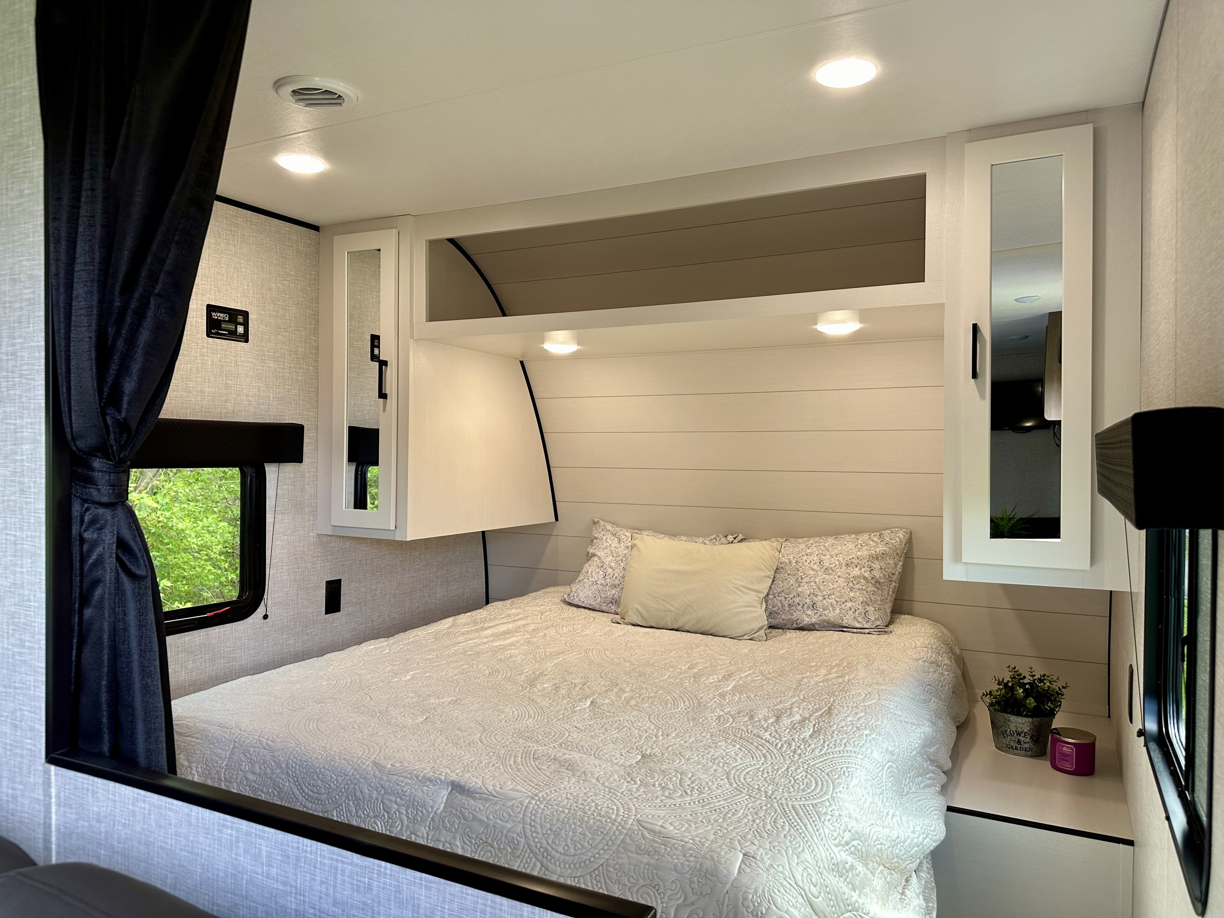 Cozy RV rental bedroom: large white bed with patterned quilt, overhead cabinets with mirrors, window with green view.. Jayco JAY FLIGHT SLX 260BH 2024