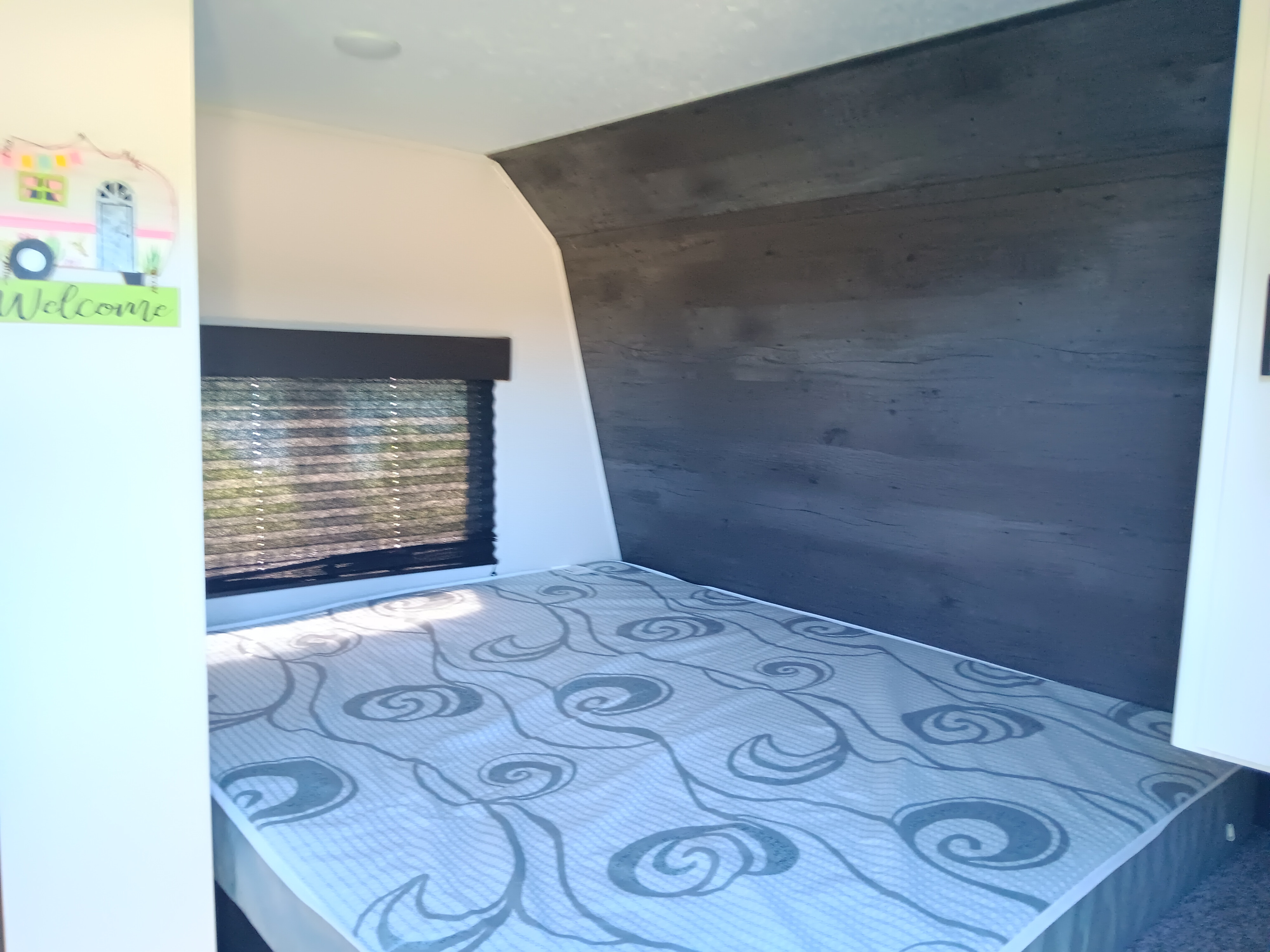 Inside this cozy rv rental, a bedroom area features a patterned bed, window with blinds, and a stylish dark wood-look accent wall.. Keystone Hideout 2021