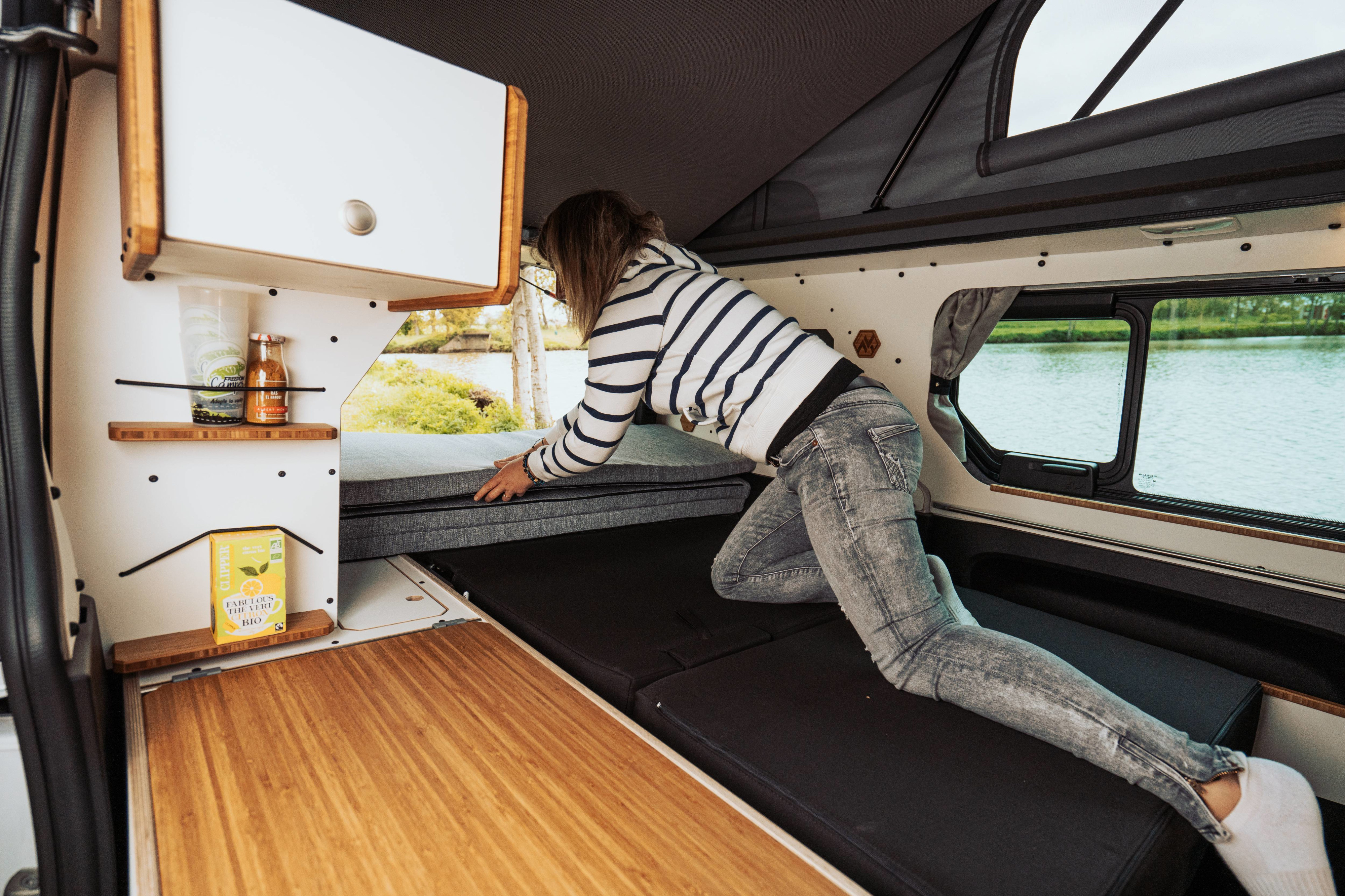 Inside this RV rental, a person arranges the modular bed. Enjoy the compact interior with shelves and a serene lake view.. Renault Trafic 2023