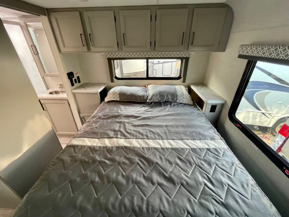 This RV rental bedroom features a cozy bed with gray bedding, overhead cabinets, and an adjoining bathroom. Perfect for your travel needs.. Thor Coleman 2023