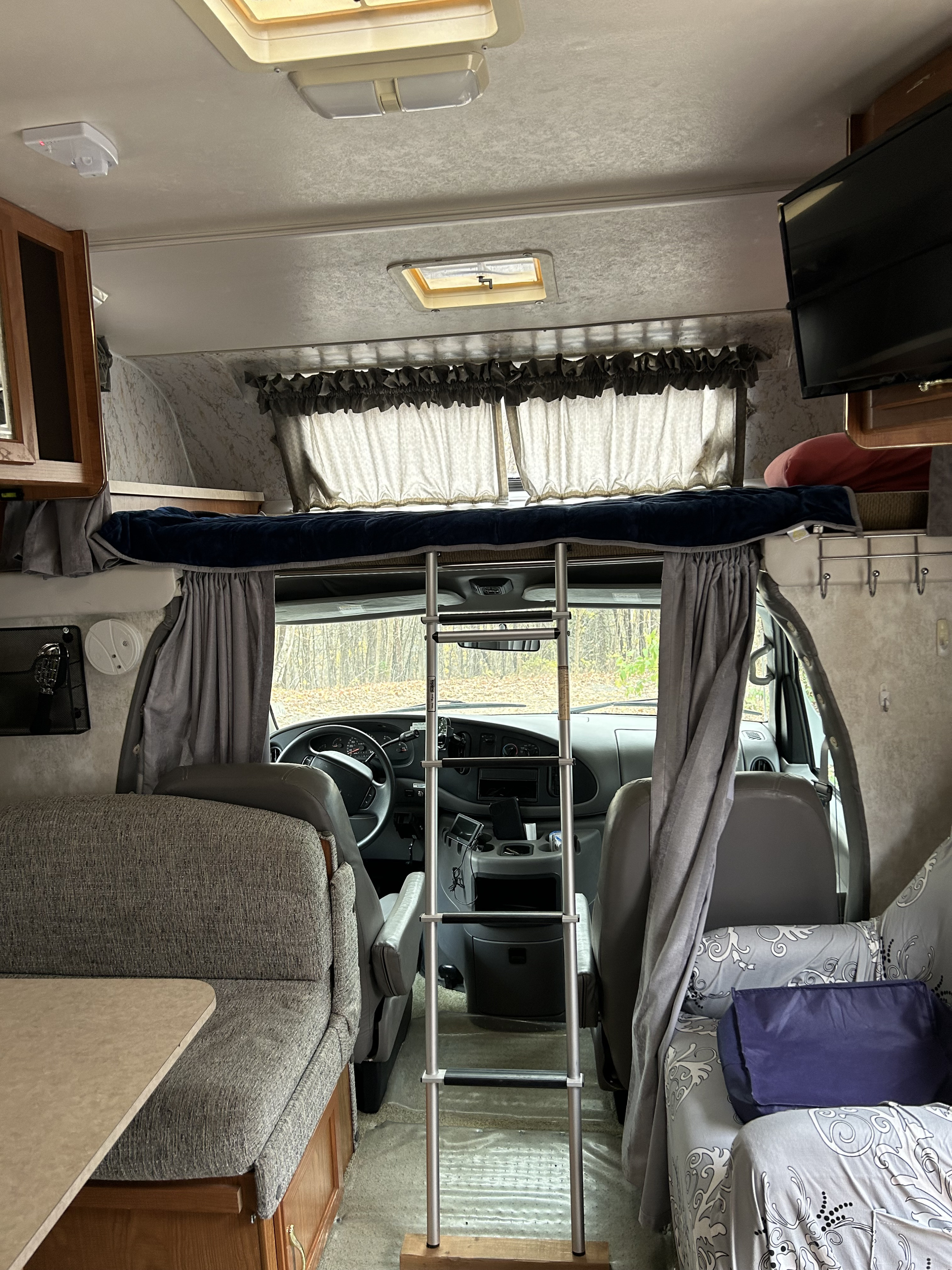 An RV rental interior shows the over-cab bunk with ladder, front cabin, and dinette area, parked with a view of trees.. Coachmen Catalina 2003