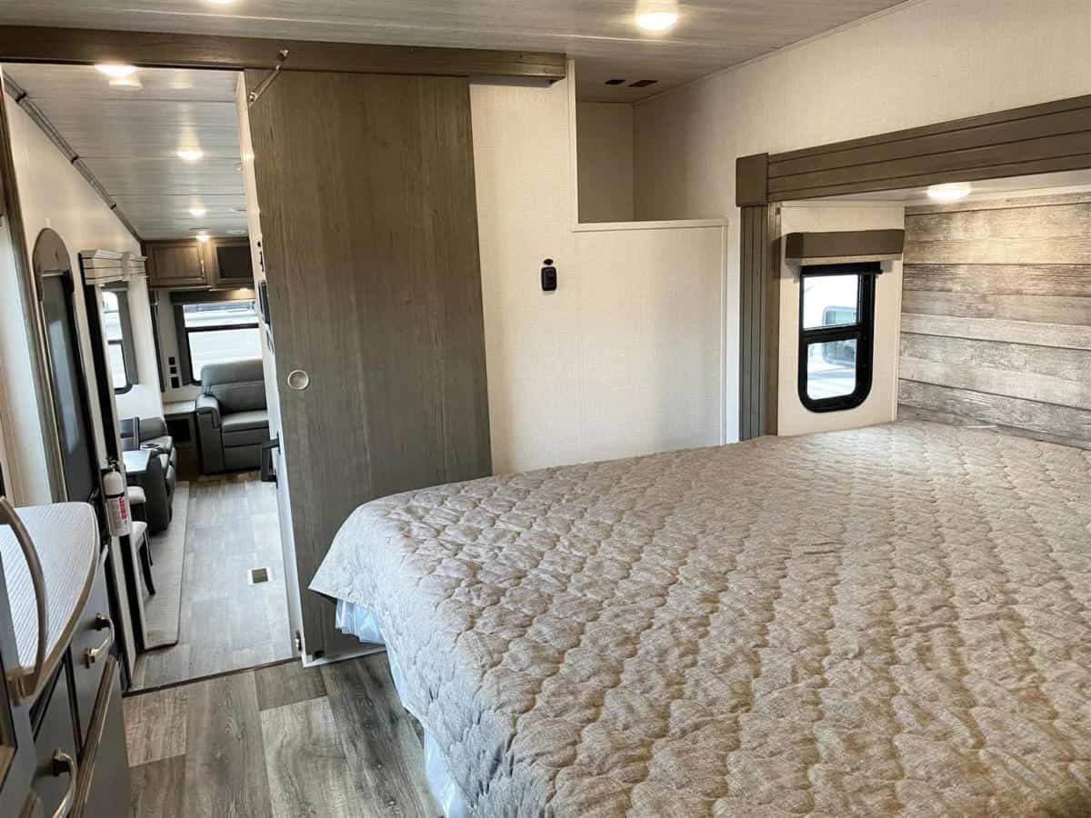 View inside an RV rental: bedroom with patterned bedding, small window, and sliding door to living area with a sofa.. Keystone Cougar 2022