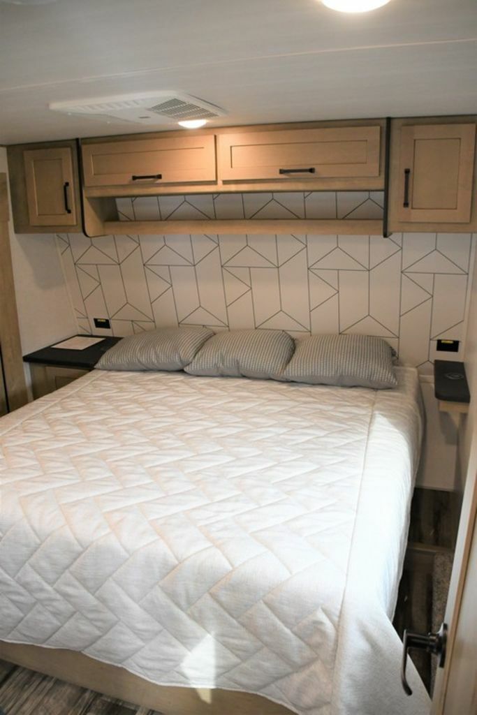 Inside an RV rental, a compact bedroom features a quilted bed, overhead storage, and modern geometric wallpaper.. Keystone Raptor 2022