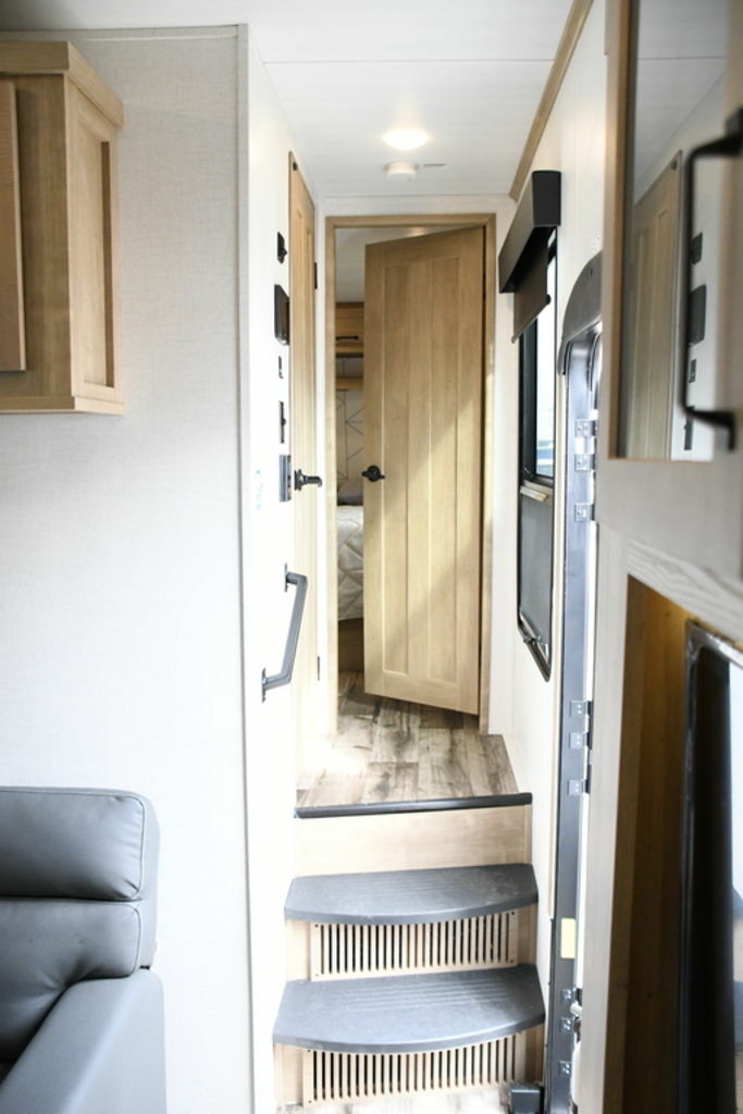 An RV rental interior: steps ascend to a hallway. A light wood door opens to a bedroom. Gray sofa visible on left.. Keystone Raptor 2022