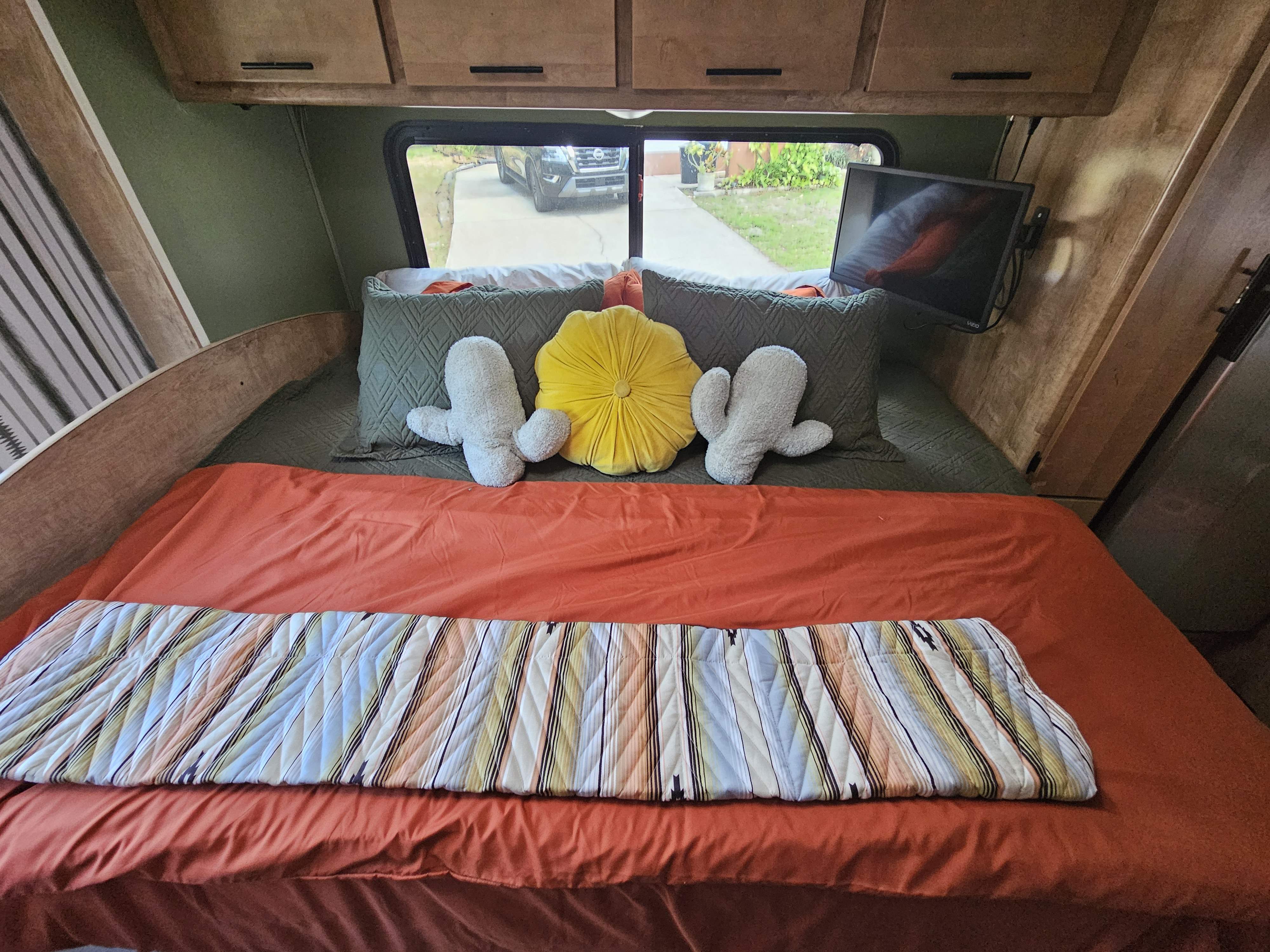Relax in this RV rental's comfy bedroom! Features orange bedding, playful pillows, a TV, and window view.. Coachmen Leprechaun 2021