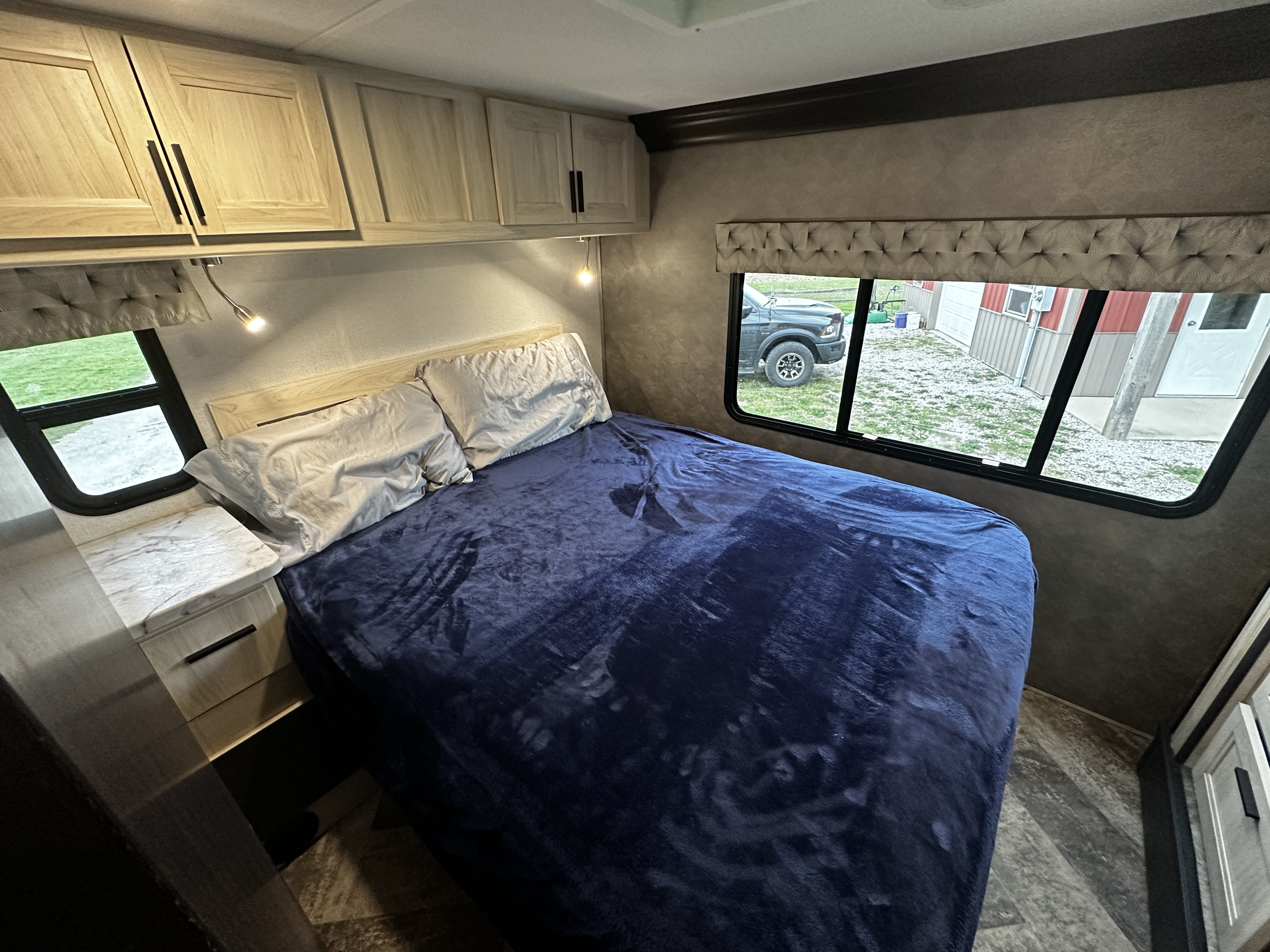 Relax in your RV rental with this bedroom's light wood cabinets, cozy blue blanket, reading lights, and window views.. Forest River Sunseeker 2020