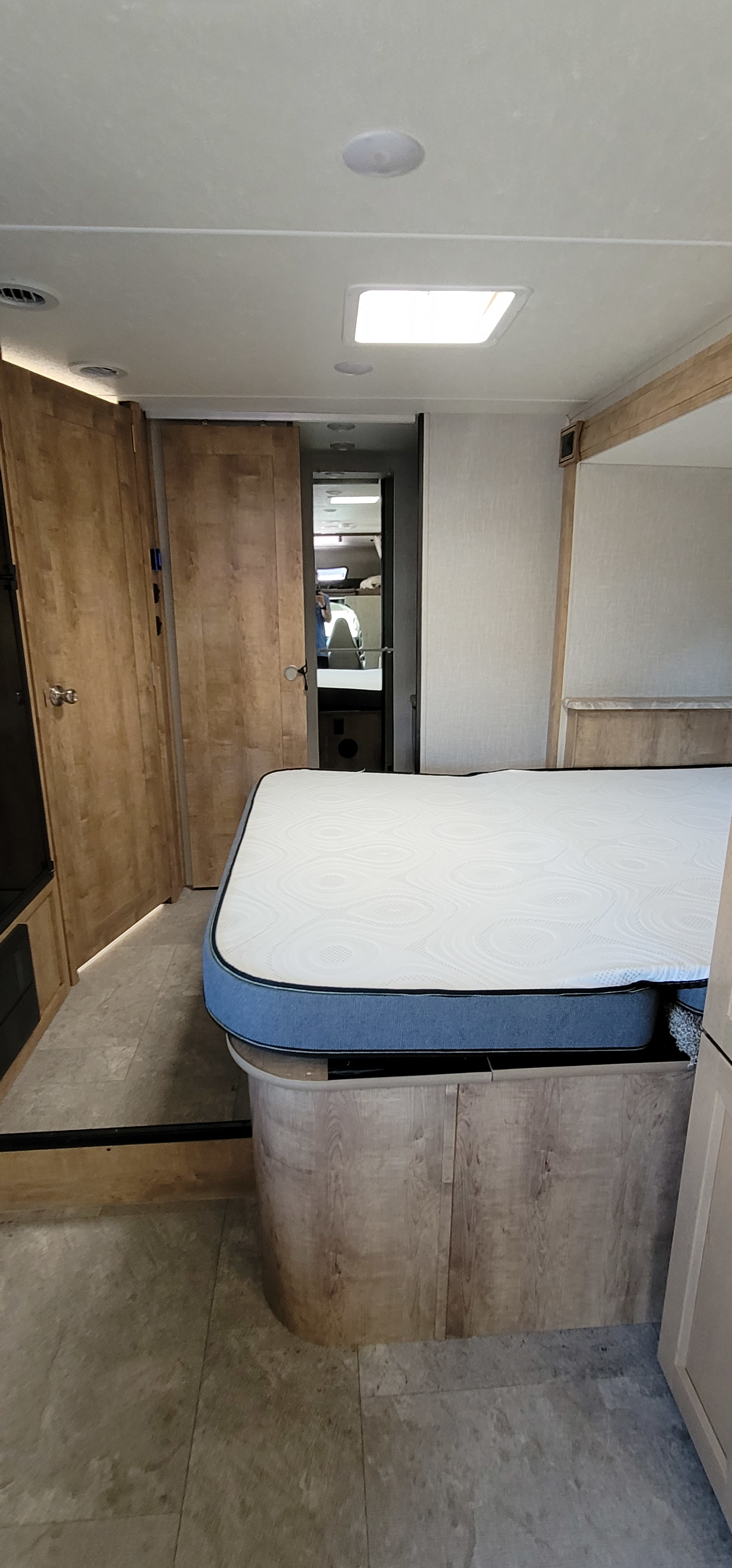 Bright RV rental bedroom showing a mattress on a wooden base, with light-toned walls and flooring, and a hallway leading back.. coachman leprachan 2020