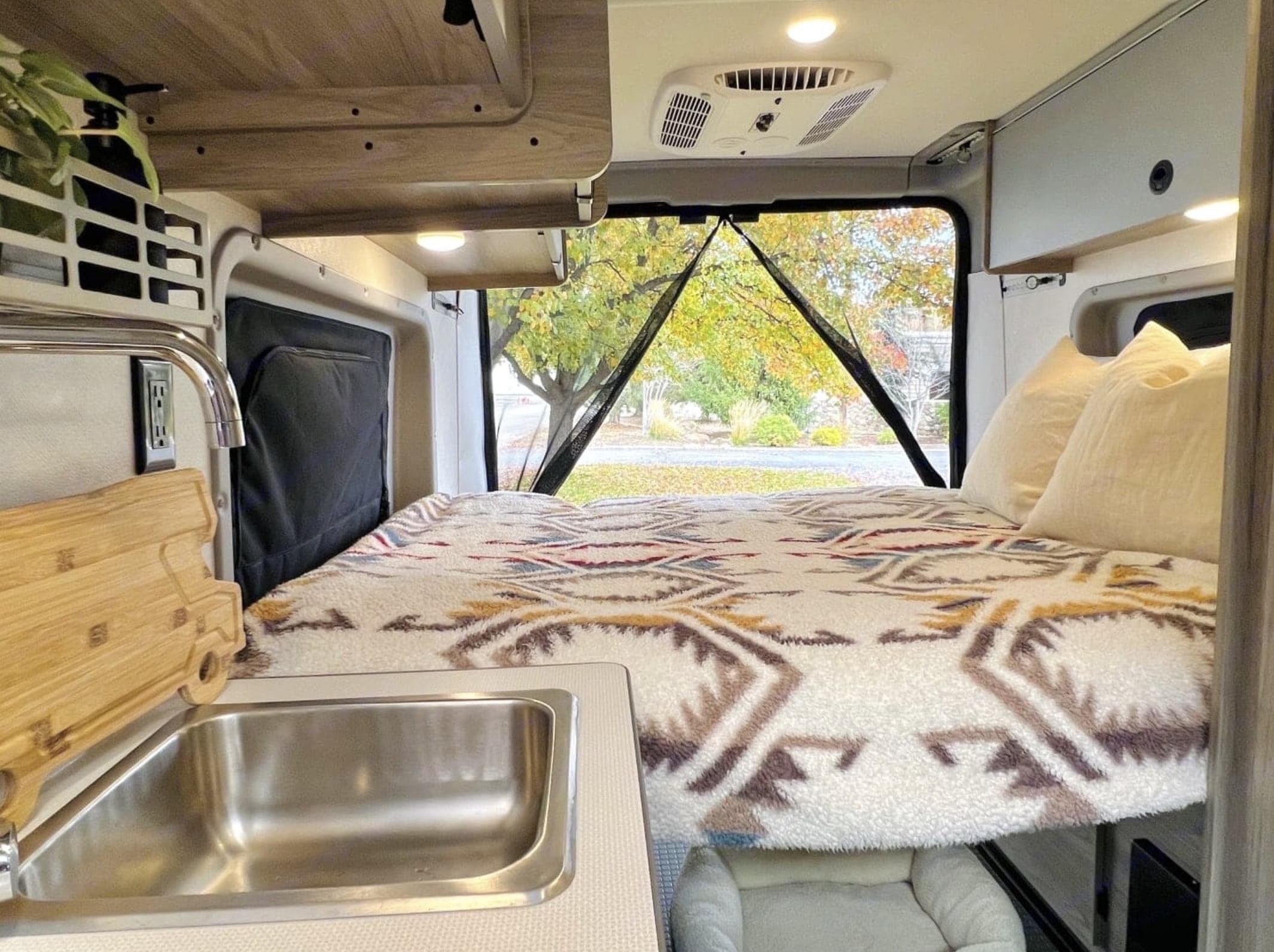 Interior of an RV rental: a cozy bed with a patterned blanket, sink, and autumn trees visible through the rear window.. Winnebago Solis 2022