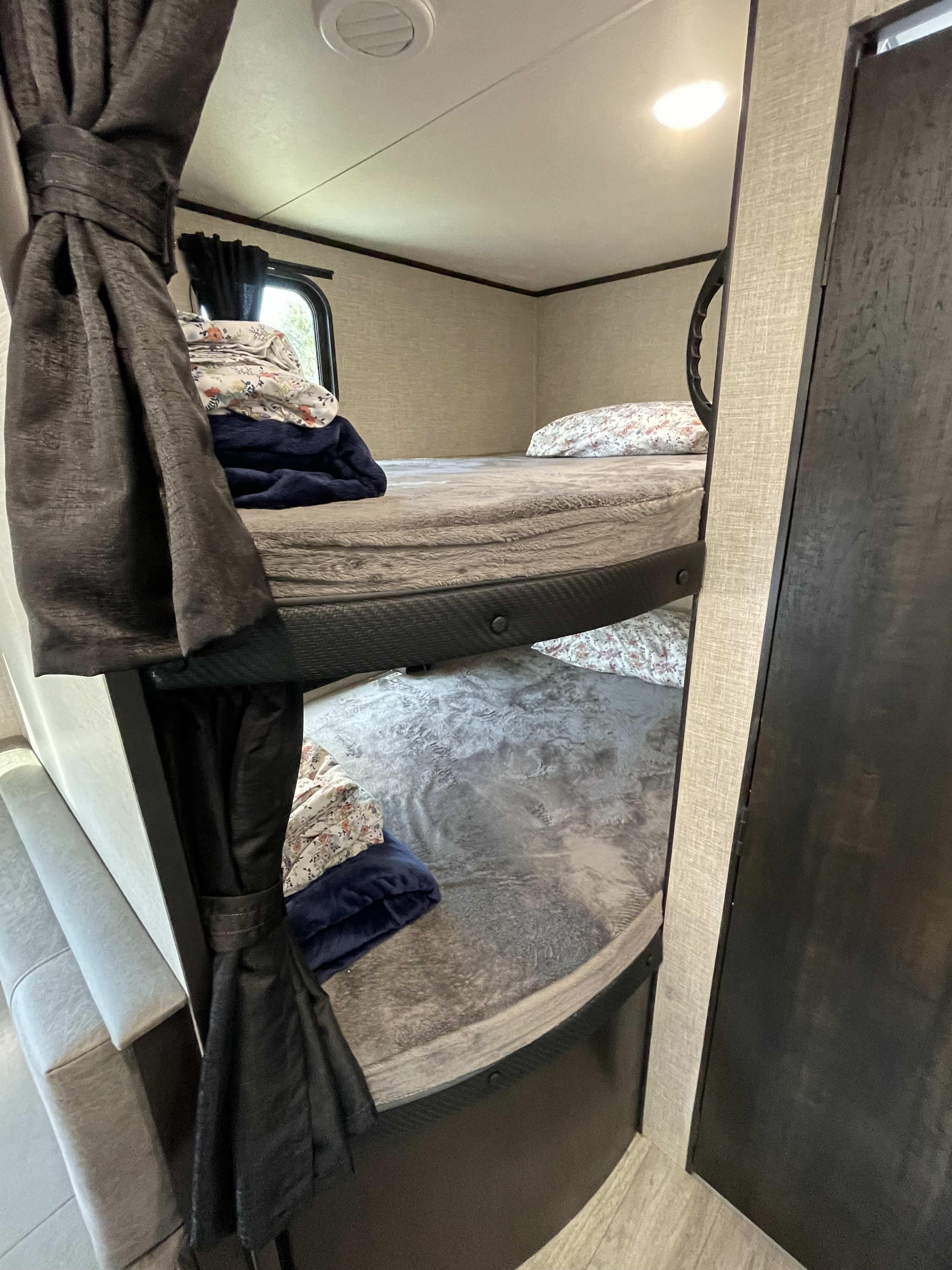 Interior view of an RV rental: two neatly made bunk beds with pillows and blankets. Dark curtains are tied back, showing a window.. Jayco Jay Flight 2022