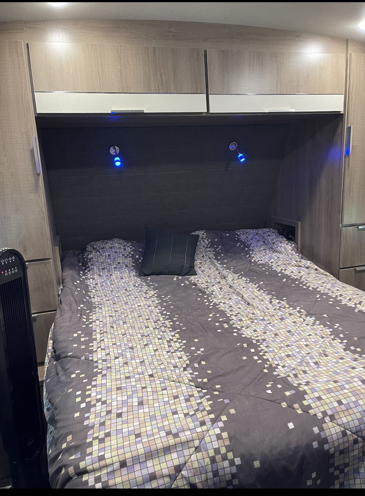RV rental bedroom: bed with mosaic comforter, built-in storage, blue LED lights, and fan on the left.. Grand Design imagine 2020