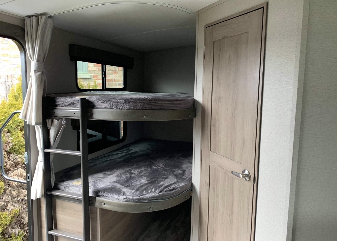 Inside an RV rental: two bunk beds with a ladder, a window view, and a wood door, offering functional sleeping space.. Grand Design imagine 2020