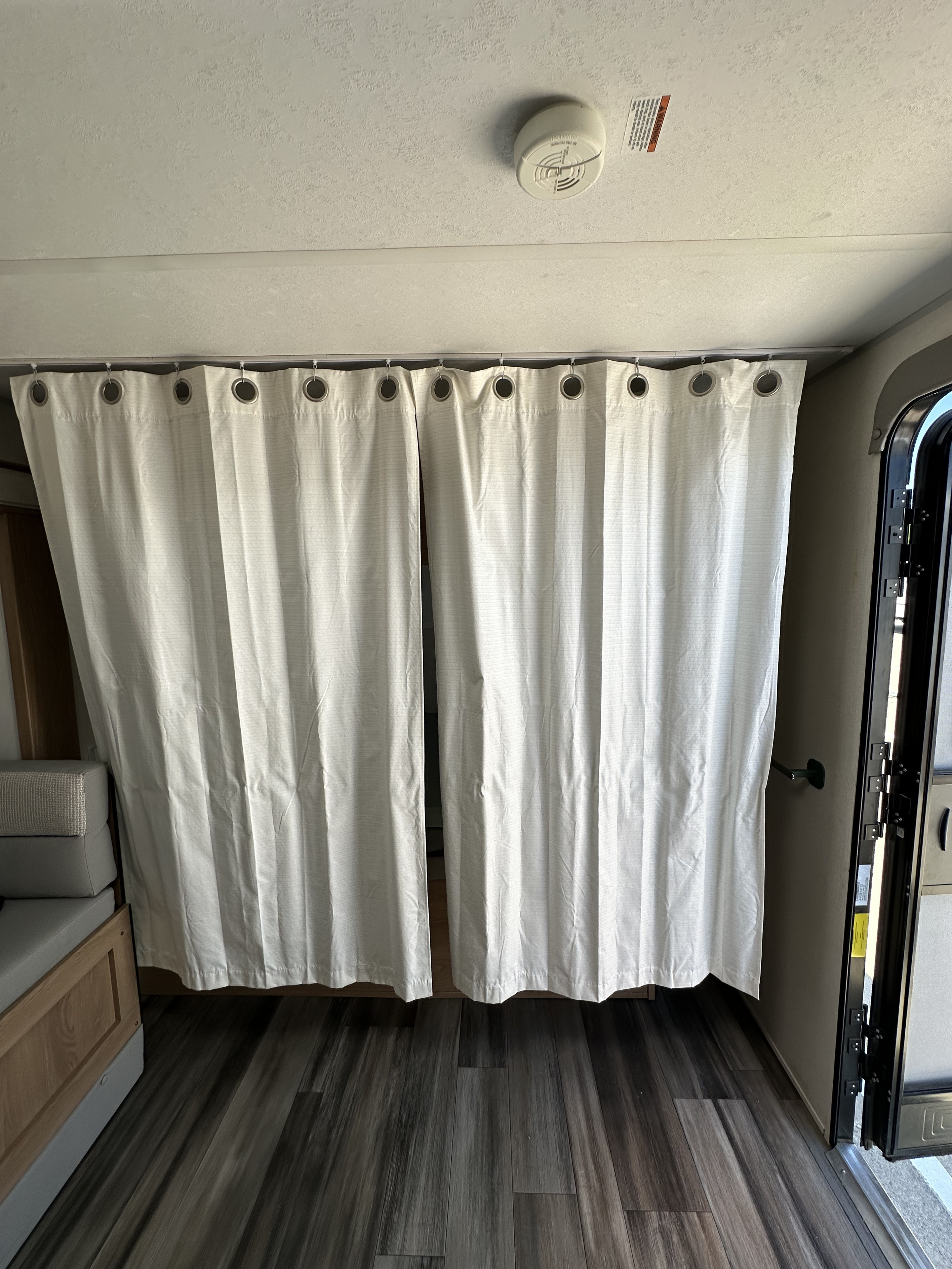 RV rental interior featuring white privacy curtains, a smoke detector, and wood-look laminate flooring, with seating and an open door.. Prime Time Avenger - 17BHS 2022