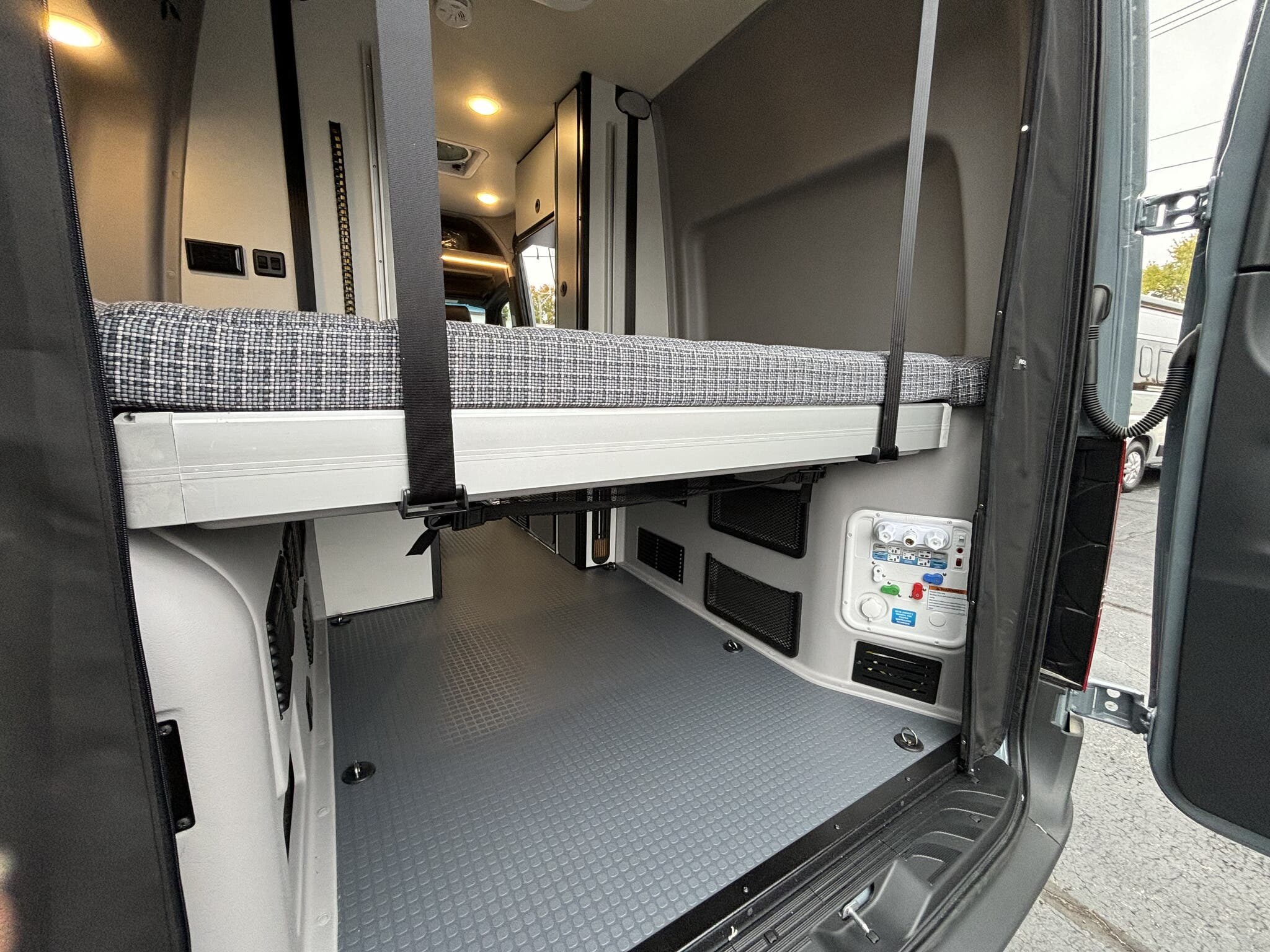 Inside an RV rental, featuring a raised bed over a spacious cargo area with a patterned floor. Utility panel on wall, rear doors open.. Winnebago Revel 2024