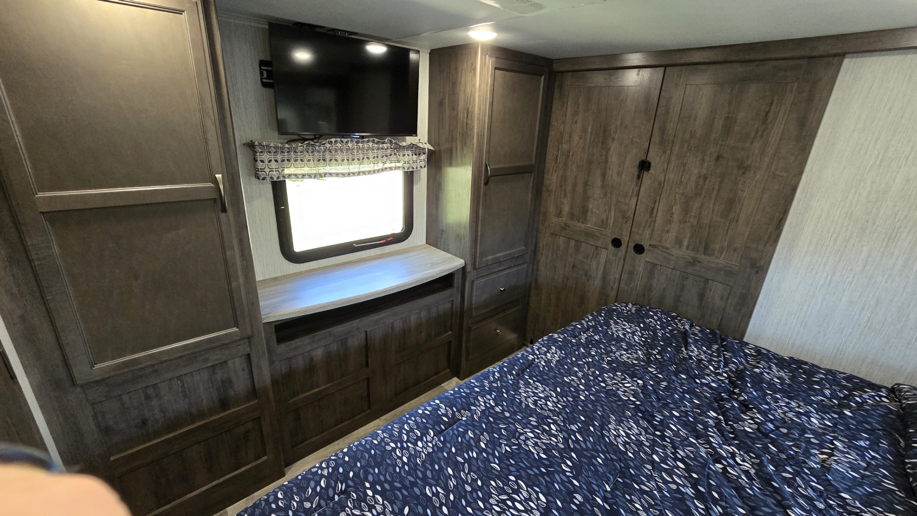 RV rental master bedroom with dark wood storage, a wall-mounted TV, and a patterned blue and white bed.. Forest River Sandpiper 2021