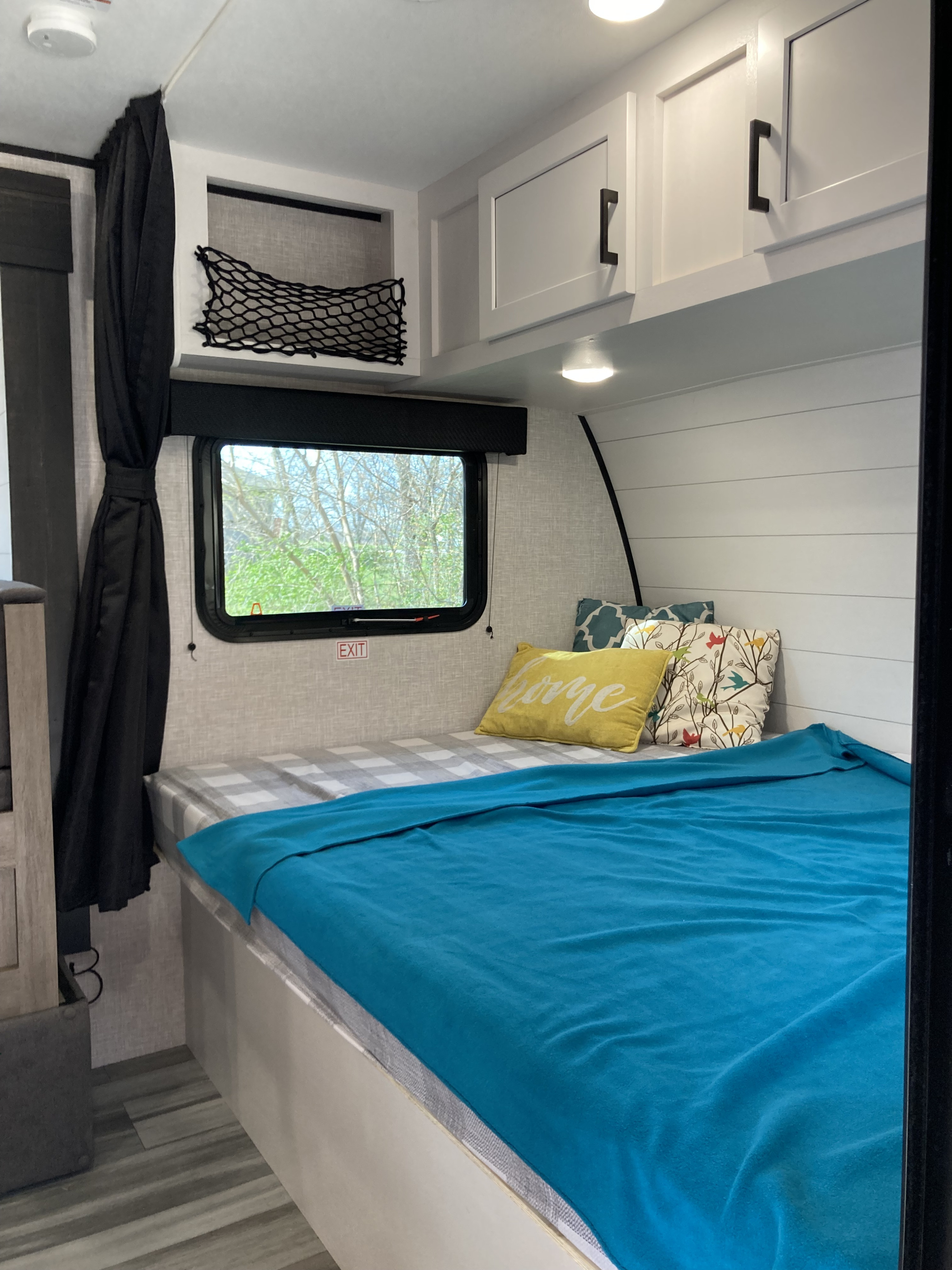 Cozy RV rental bedroom with a comfortable bed, blue blanket, and decorative pillows. Features overhead cabinets and a bright window view.. Jayco Jay flight 184bs 2024