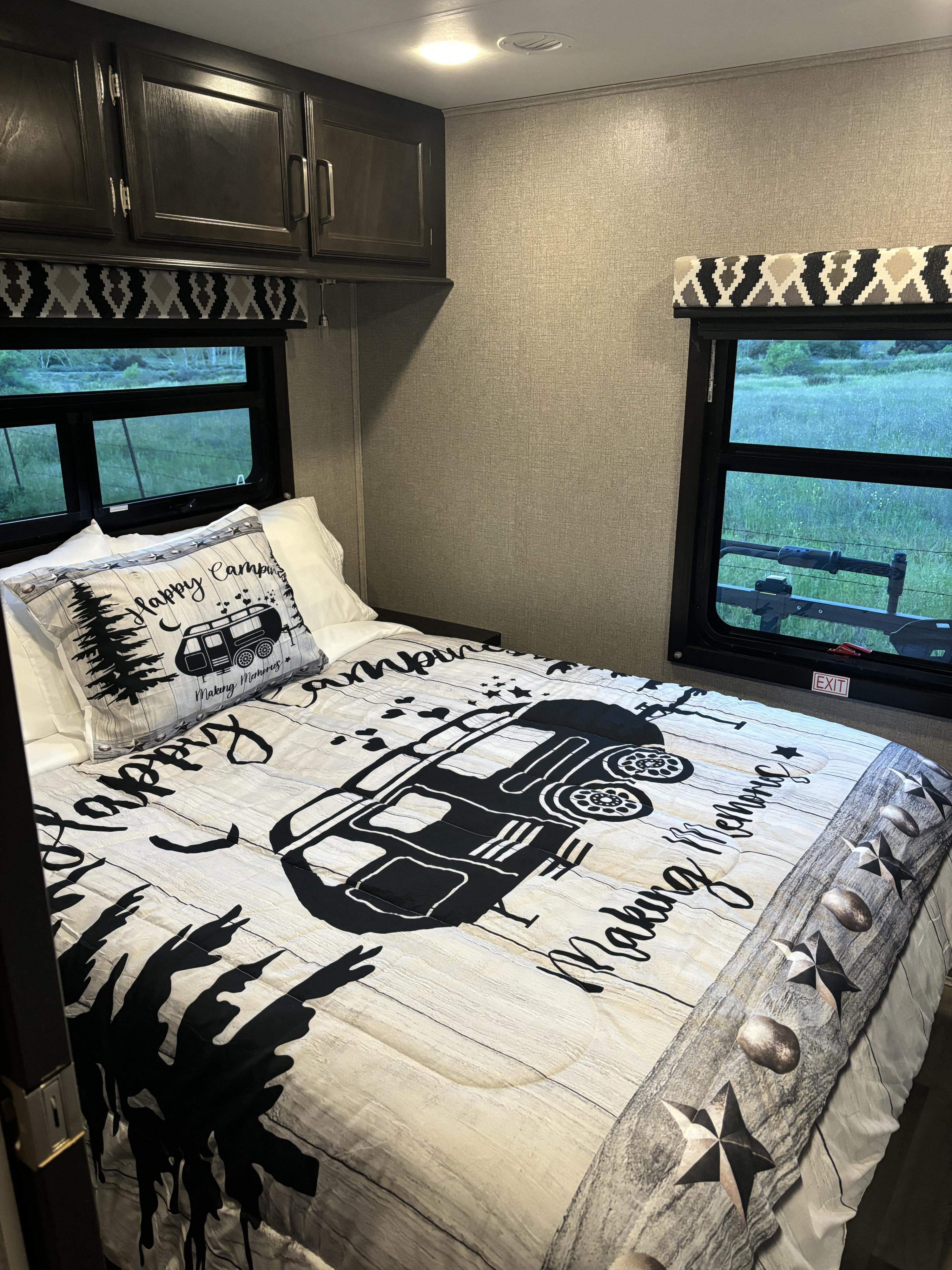 Cozy RV rental bedroom with "Happy Camper" bedding, dark cabinets, and scenic windows showcasing lush greenery.. Entegra Coach Odyssey 2021