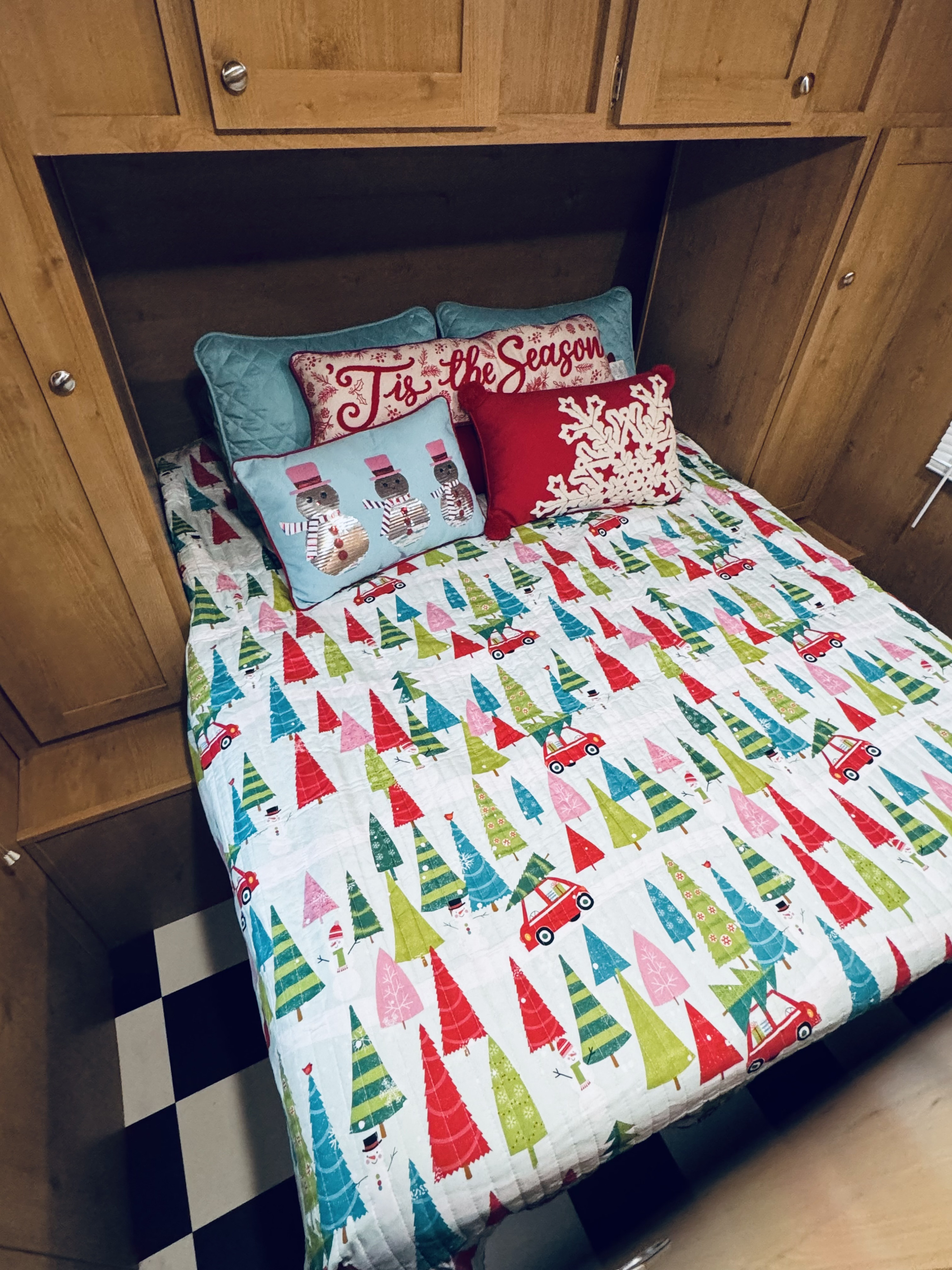Cozy RV rental bed area featuring festive holiday tree quilt, snowman, and 'Tis the Season' pillows amidst wood cabinetry.. Riverside RV Retro 2021