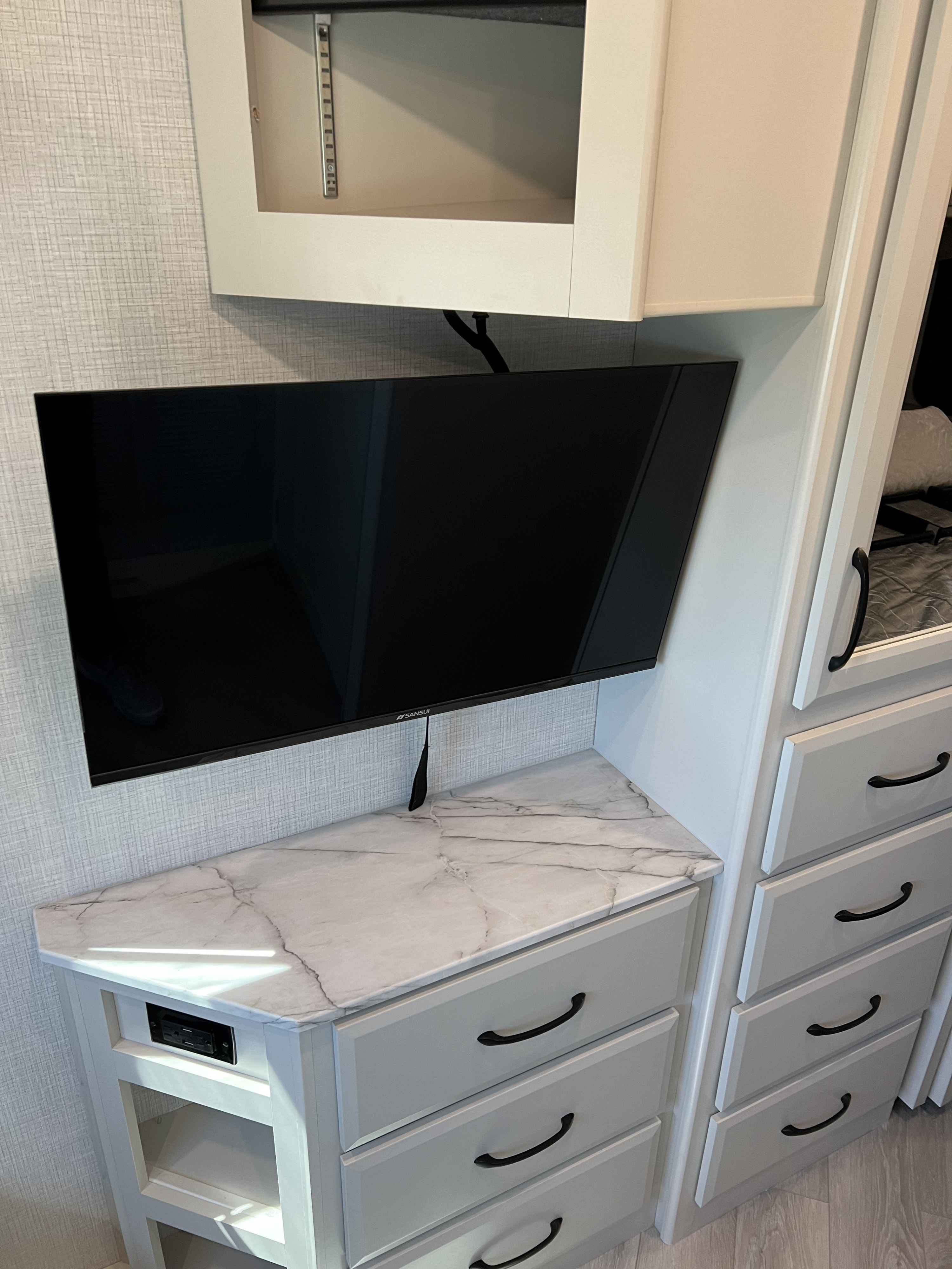RV rental interior: wall-mounted TV, white bedroom cabinets with black pulls, and a marble-look counter, offering smart storage.. Fleetwood Fortis 2024