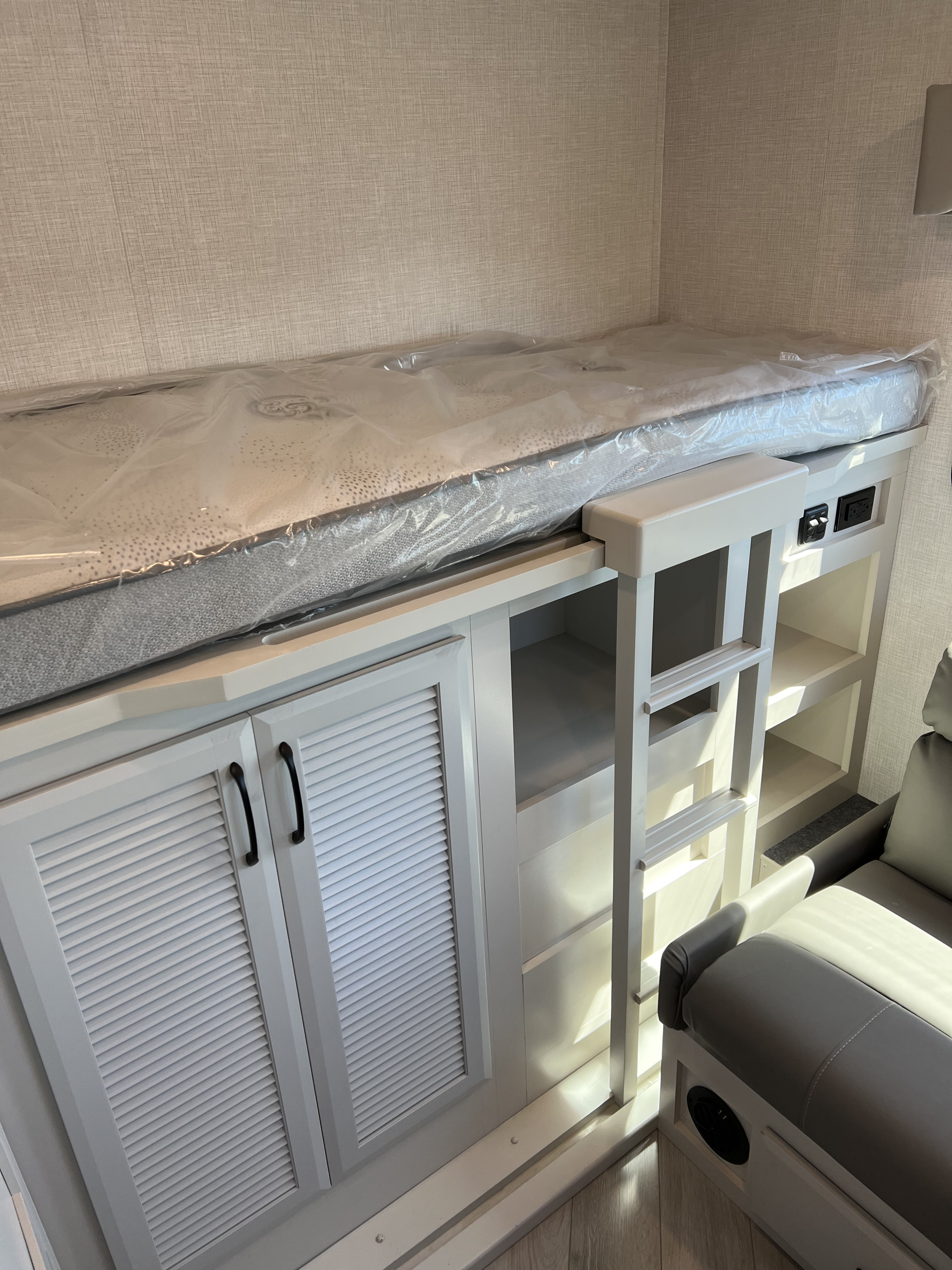 RV rental bunk bed setup: wrapped mattress, white storage cabinets with louvered doors, ladder, outlets, & gray seating.. Fleetwood Fortis 2024