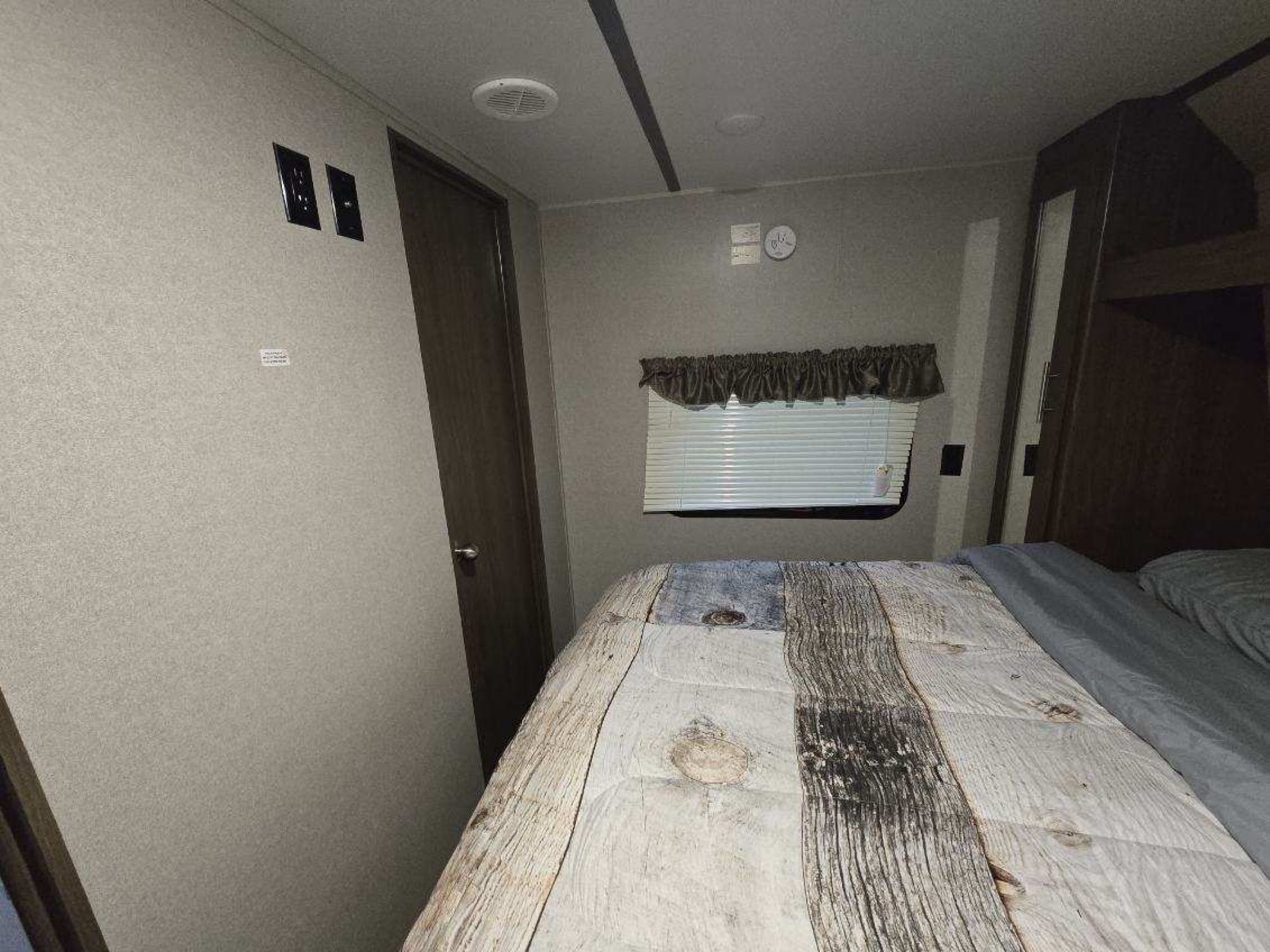 Inside an RV rental, a compact bedroom features a bed with a unique wood plank design comforter, window, and built-in storage.. Keystone RV Hideout Luxury 2019