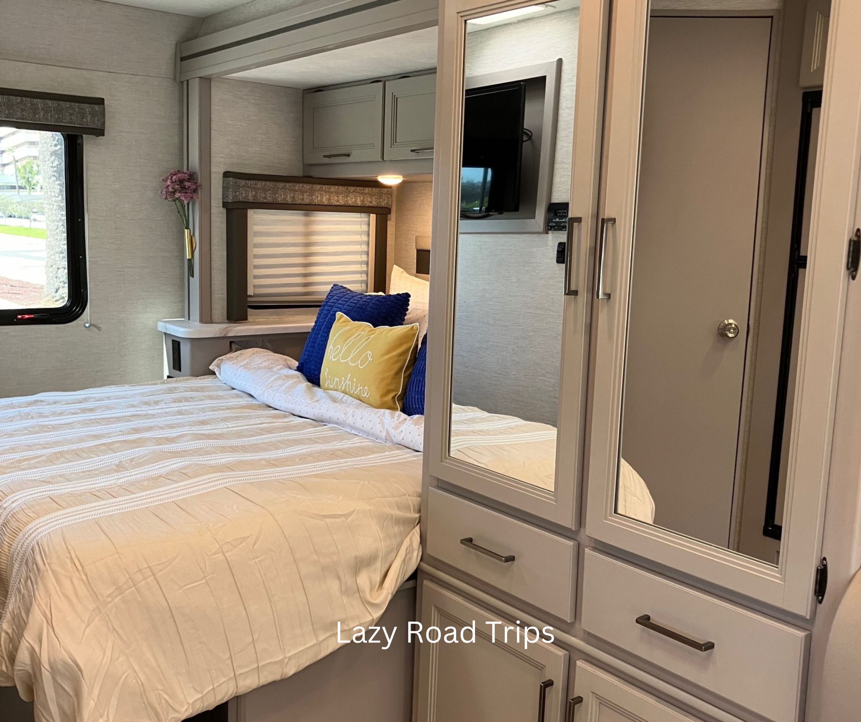 Bright RV rental bedroom: striped bedding, decorative pillows, window view, and mirrored storage.. Thor Chateau 2024