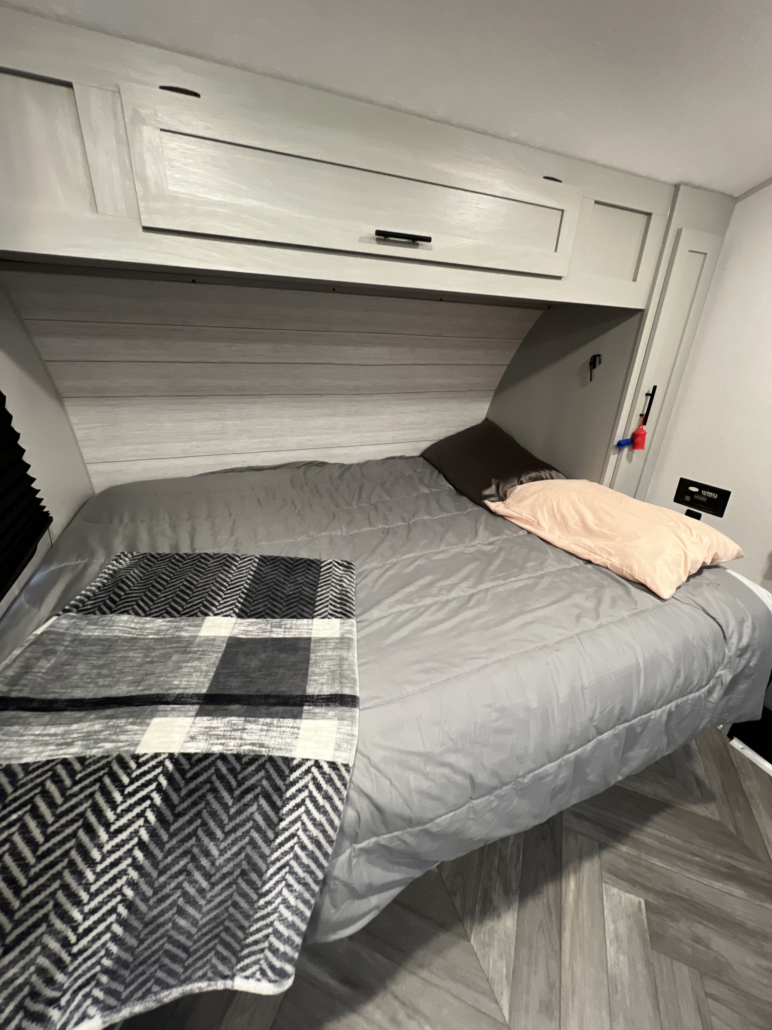 Cozy RV rental bedroom with a grey bed, plaid blanket, pillows, overhead cabinets, and wood-patterned floor.. Forest River Wildwood 2022