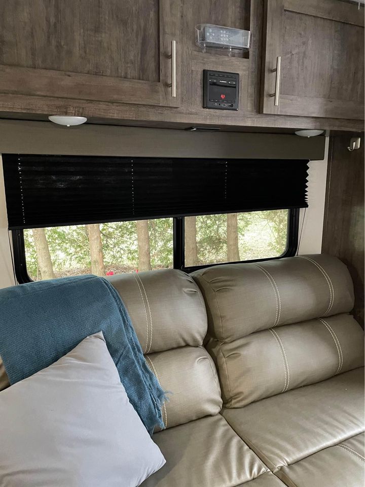 Inside an RV rental featuring wood cabinets, black blinds, a control panel, and a cozy tan sofa with a blue blanket. Trees visible outside.. Riverside RV Retro 2022