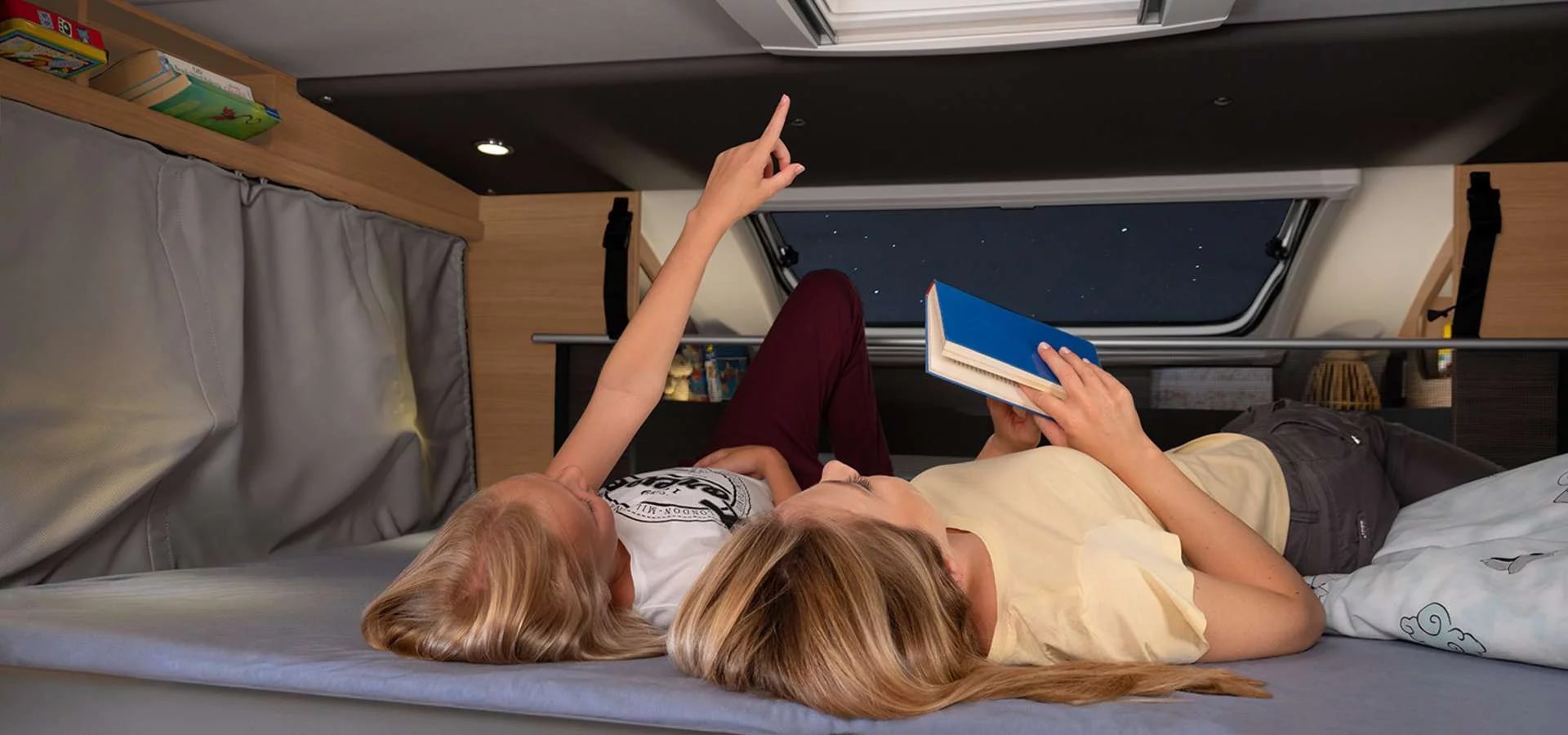 RV rental adventure: Two women lie in a bunk, one points to stars outside, the other reads a book. Cozy night.. Knaus Live Wave 650 MG 2024