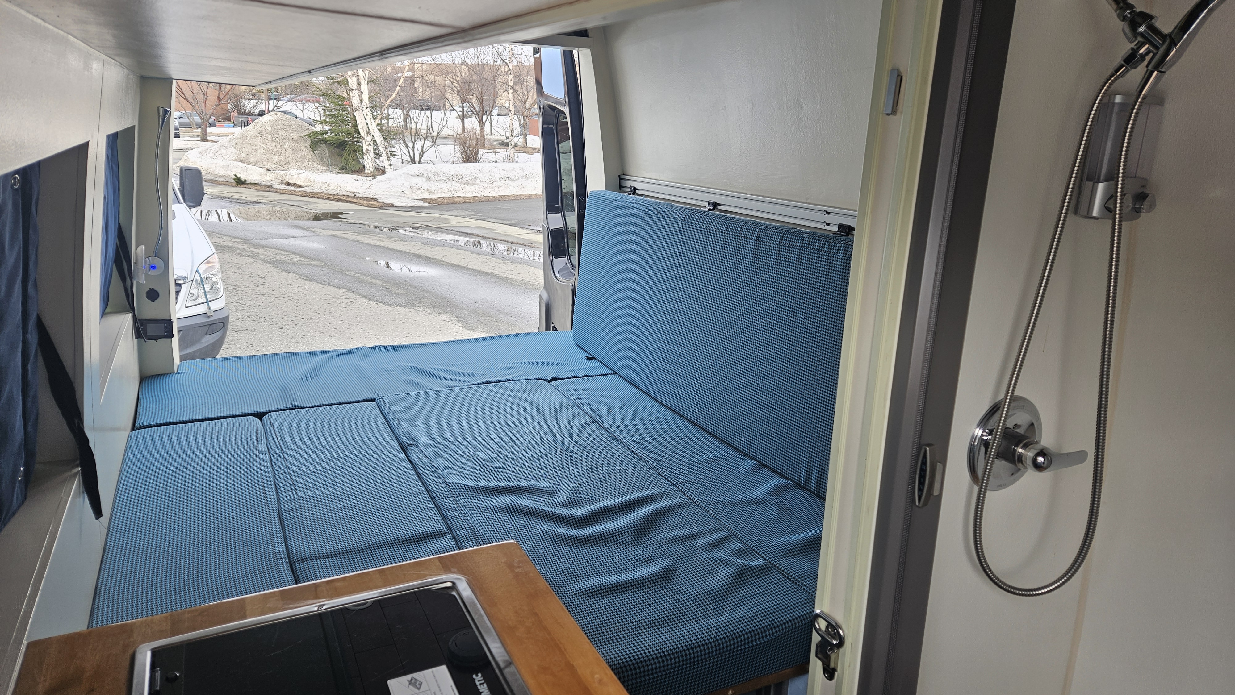Cozy RV rental interior: versatile bed, compact kitchen, and shower setup. View of snowy road outside.. Mercedes-Benz Sprinter 2015
