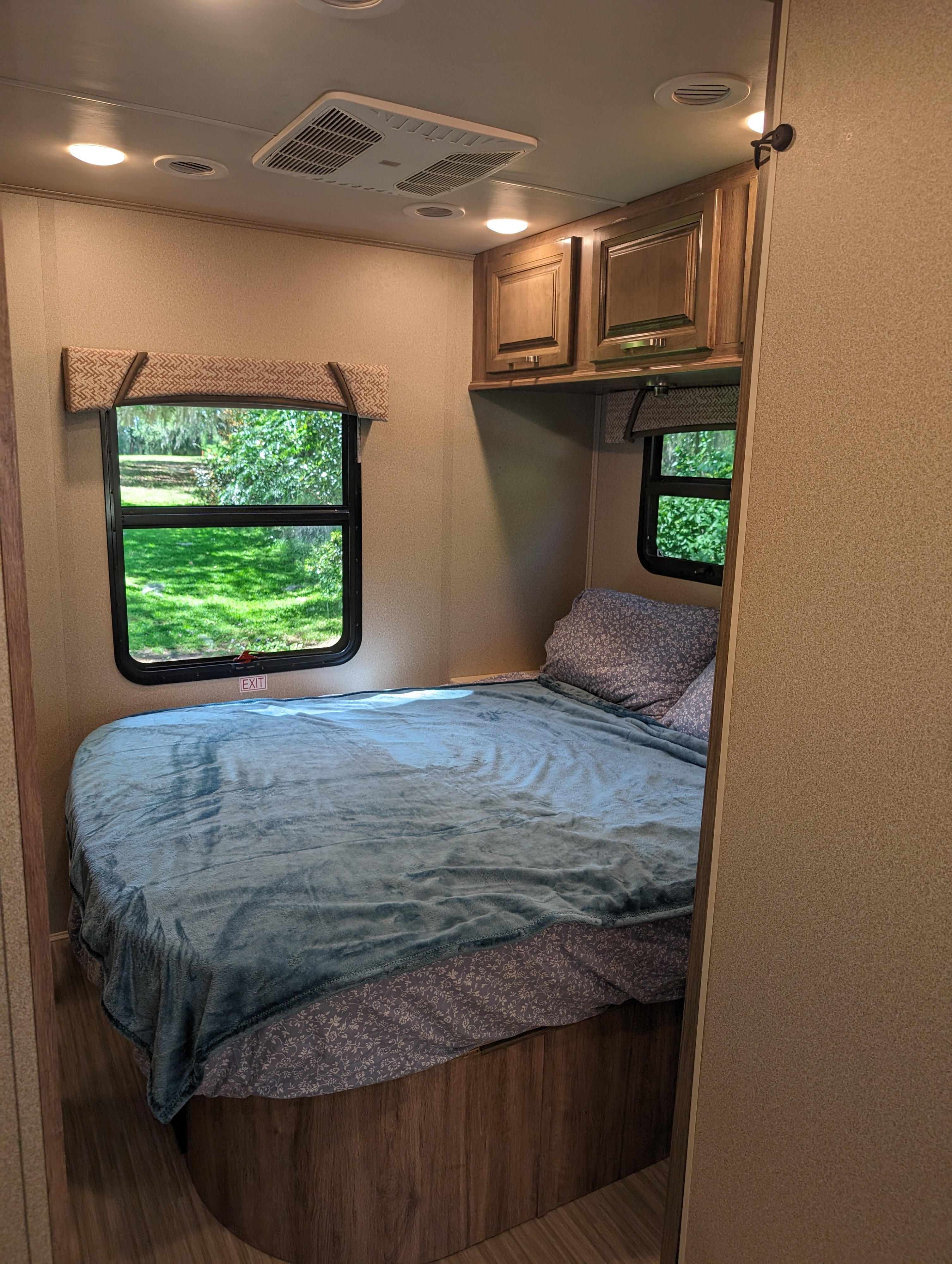 Cozy RV rental bedroom features a comfy bed with blue blanket, scenic window views, and convenient overhead storage.. Jayco Greyhawk 2019