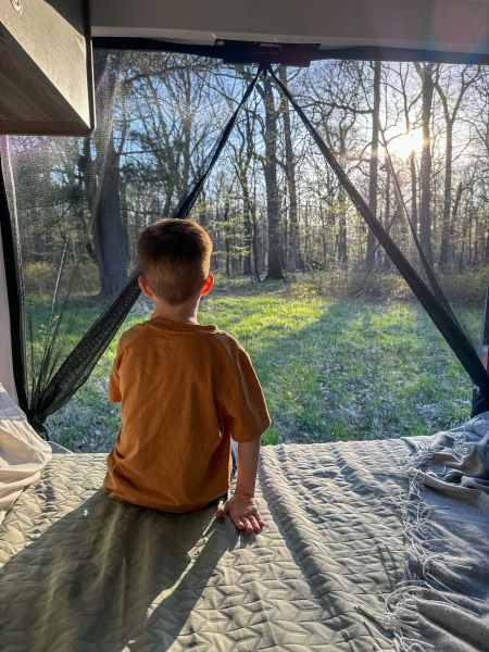 Experience nature from your RV rental! Child gazing at a sunlit forest view from the cozy bed. Ideal family adventure.. Winnebago Solis 2023