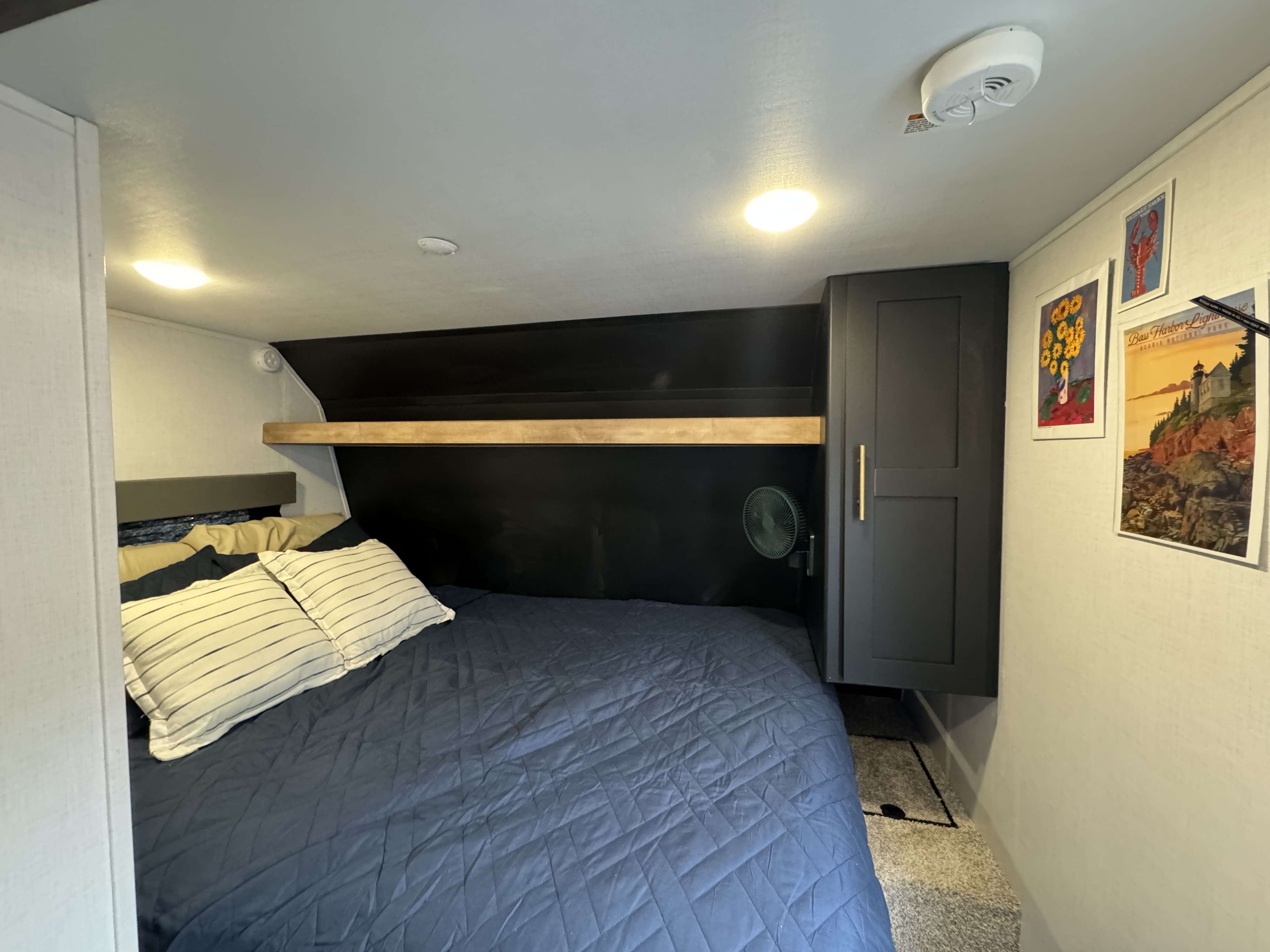 RV rental bedroom with blue bedding, dark accent wall, a mounted fan, wooden shelf, and a dark grey storage cabinet.. Keystone RV Springdale 2023