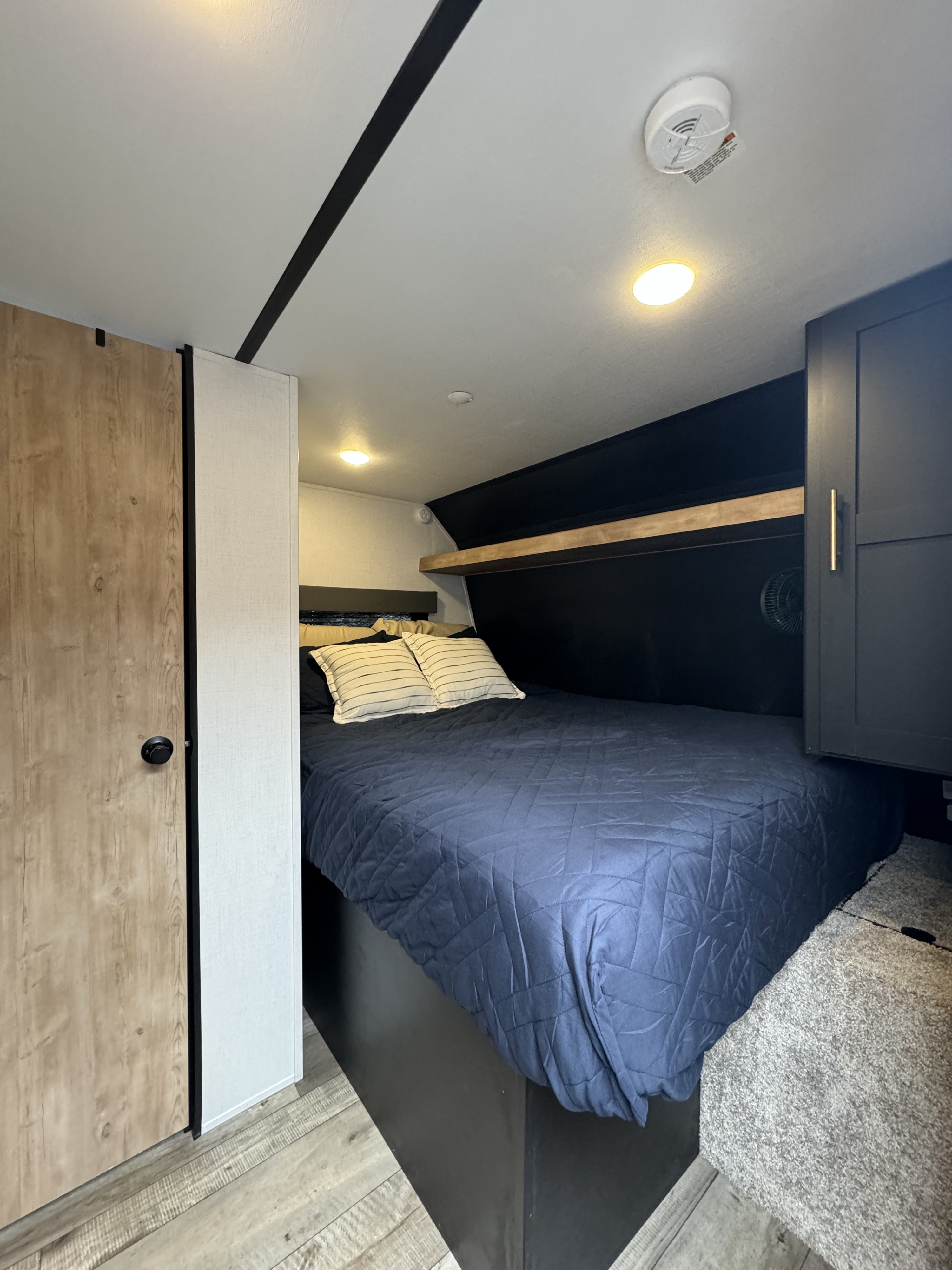 Cozy RV rental bedroom with a dark blue bed, wood accents, built-in storage, and a ceiling-mounted smoke detector.. Keystone RV Springdale 2023