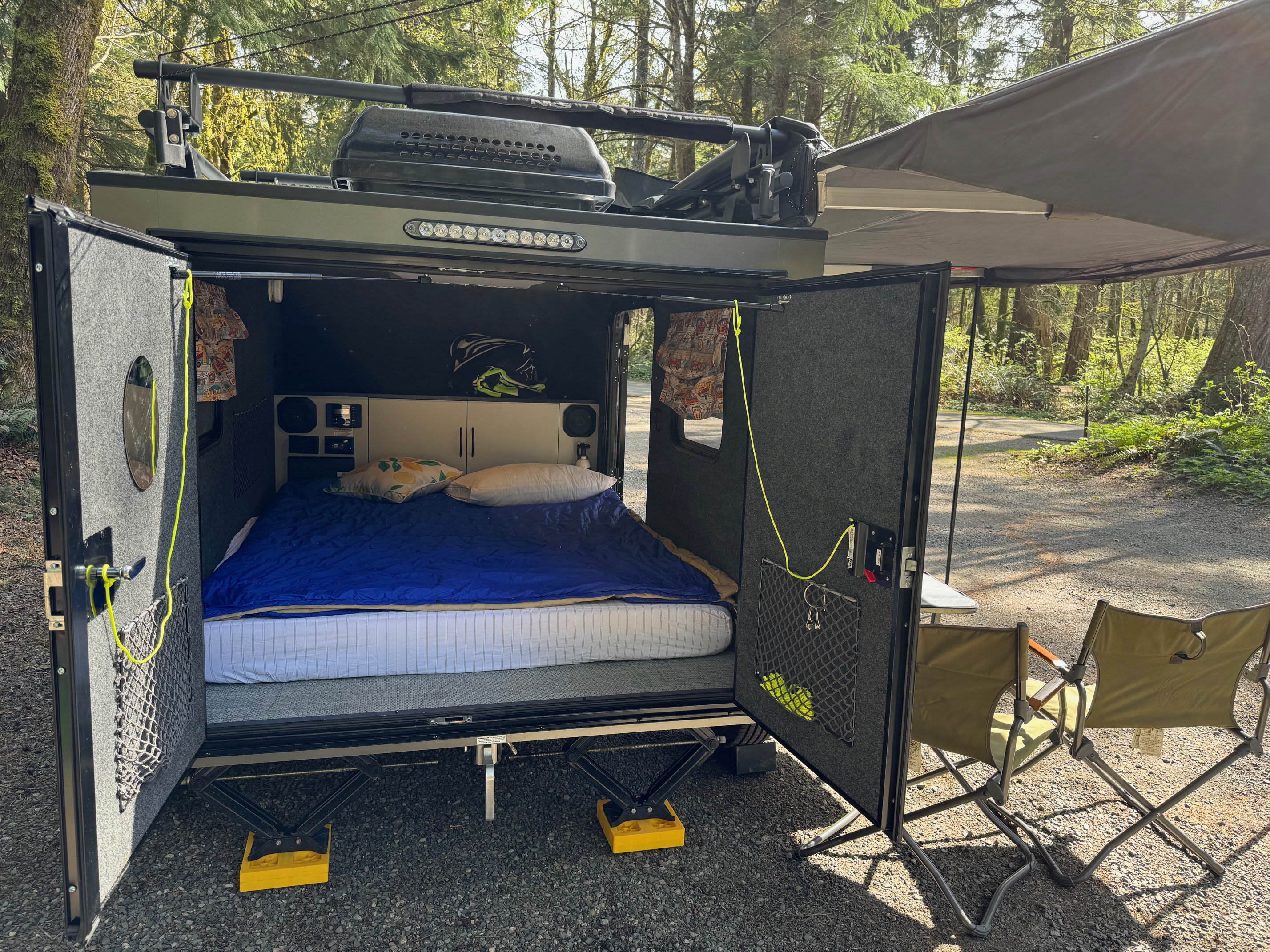Compact RV rental: A cozy camper with a bed, an extended awning, and outdoor chairs at a serene, woodsy campsite.. InTech RV InTech RV Trailer 2021