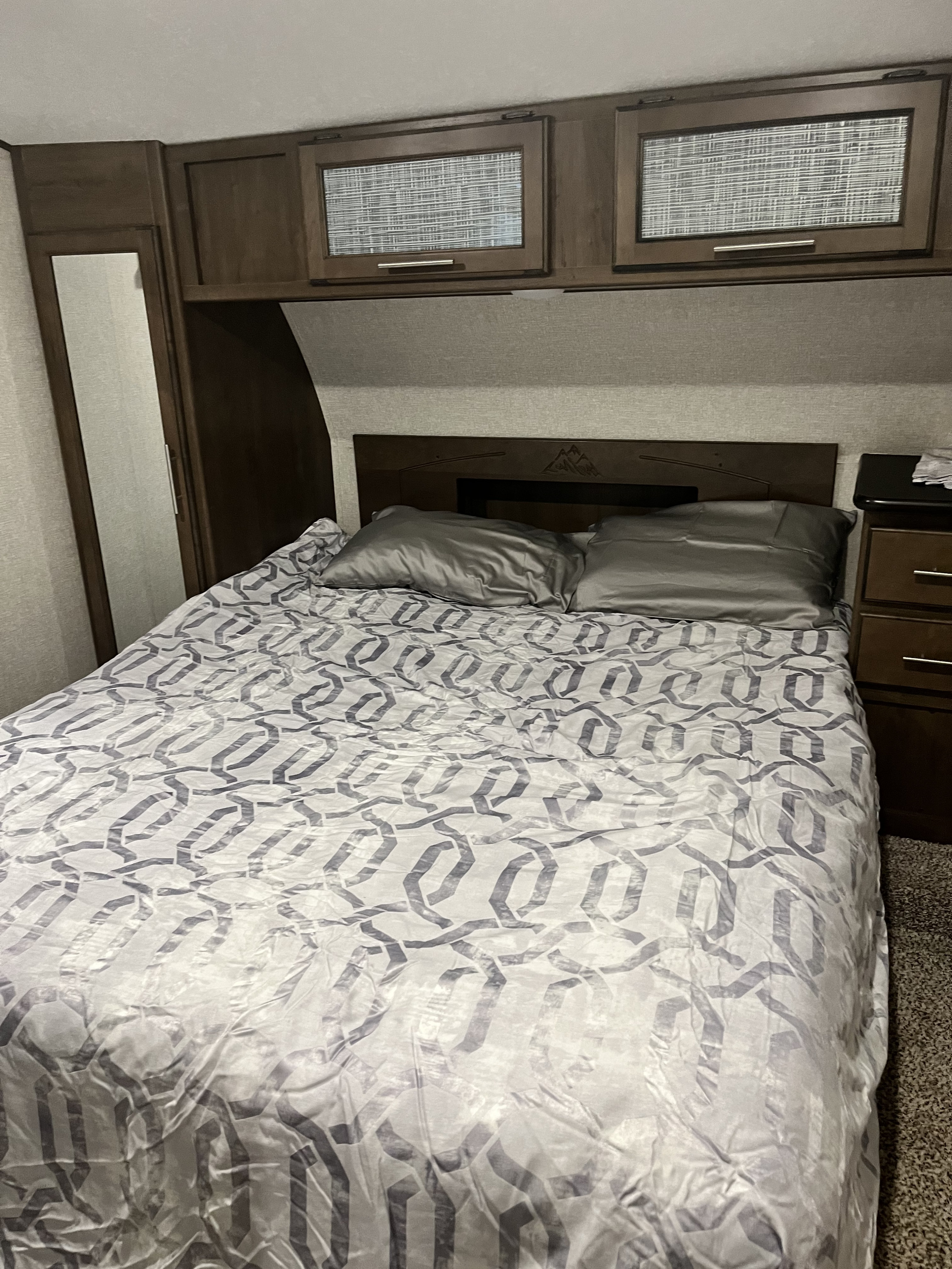 Cozy RV rental bedroom features a comfy bed with patterned comforter, grey pillows, wood cabinets, and mirror.. Keystone RV Cougar 2018