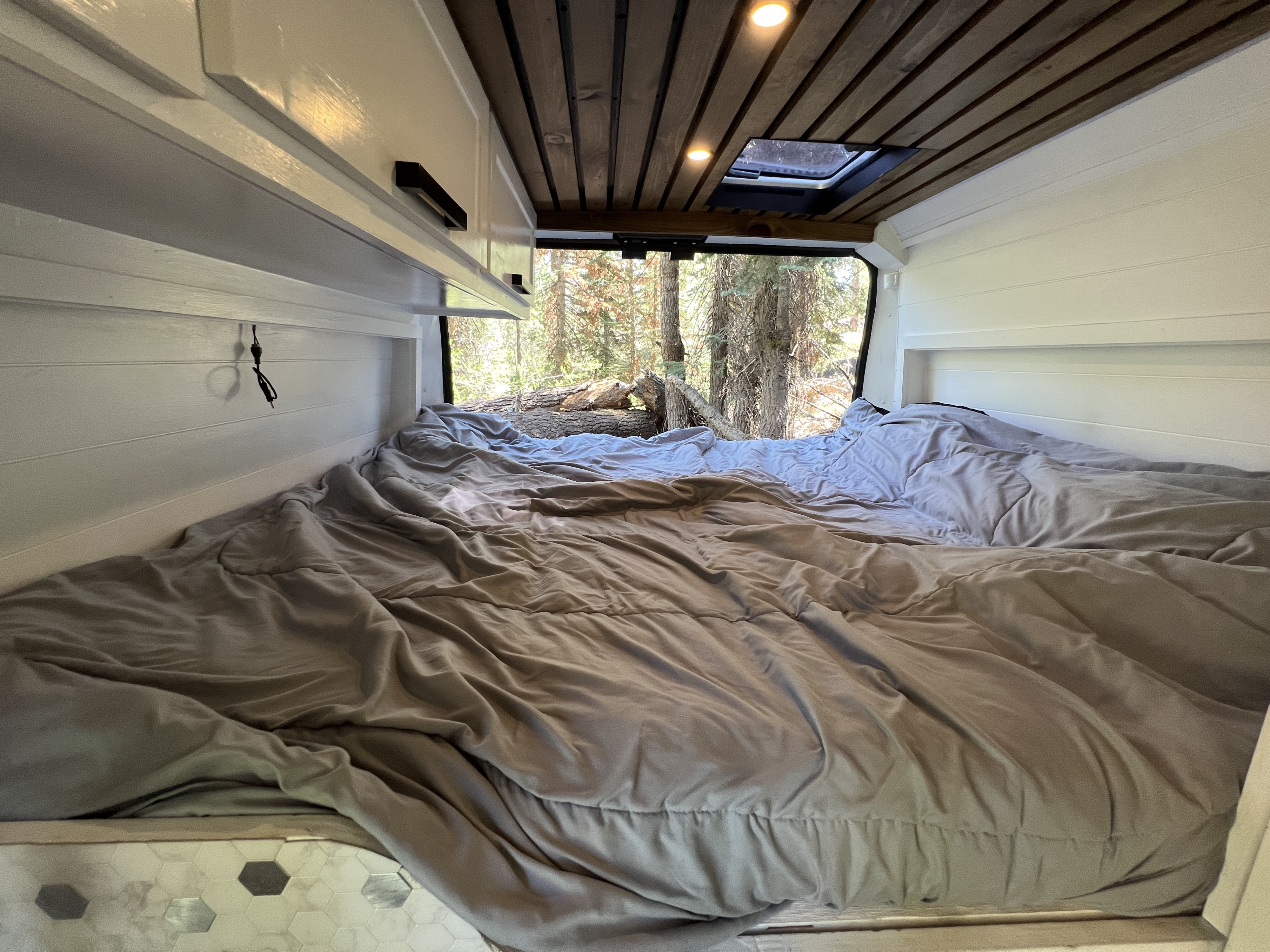 Cozy RV rental bed with white shiplap walls, wooden ceiling, and a scenic forest view from the back window.. Dodge Sprinter Van 2019